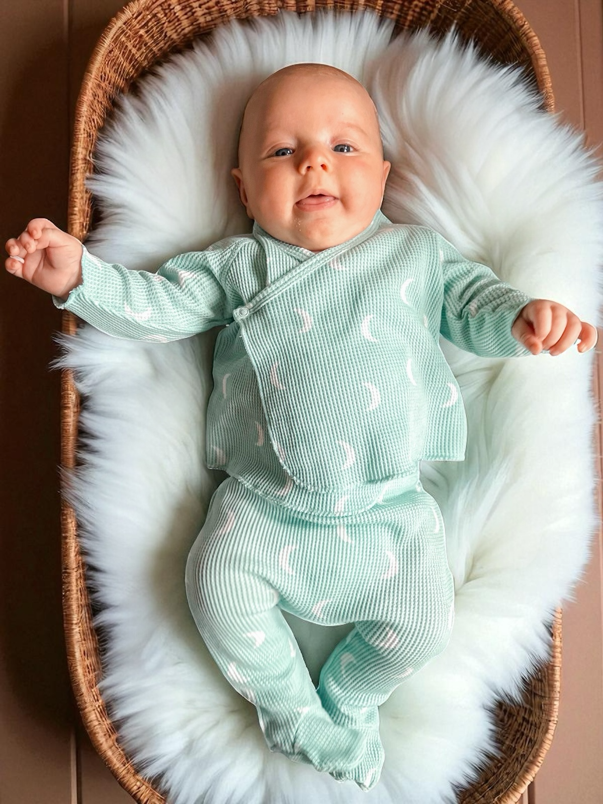 Smiling baby in mint green outfit lying on fluffy blanket in a woven basket.