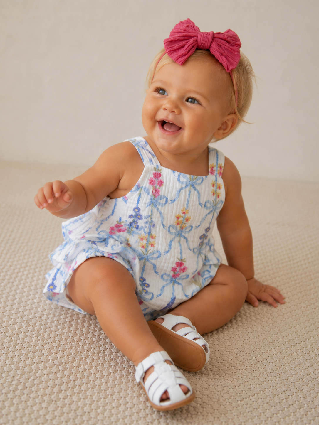 Smiling toddler in floral outfit and pink headband sitting on a textured surface.