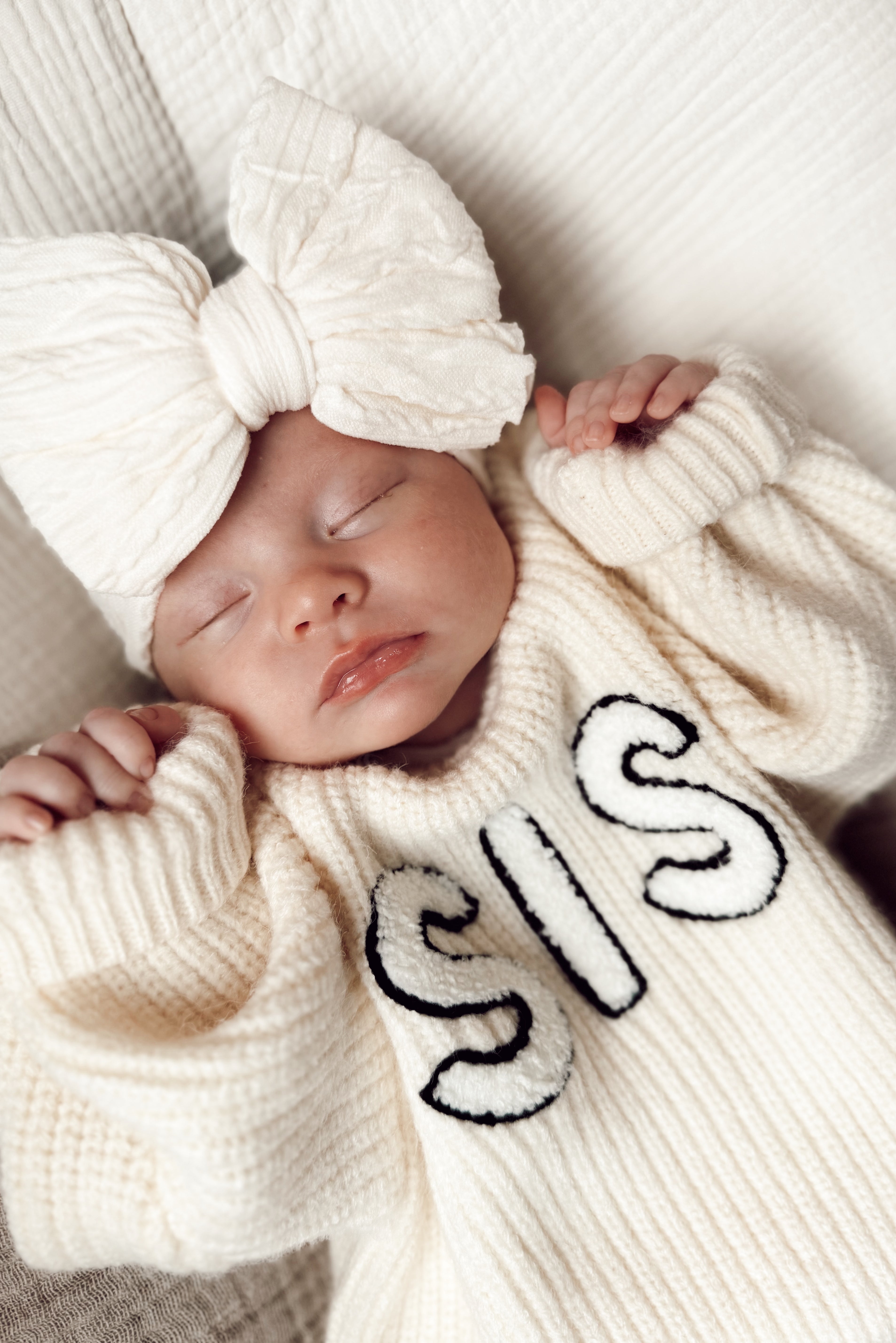 Sleepy baby wearing a cozy cream sweater with "SIS" text and a large bow headband.