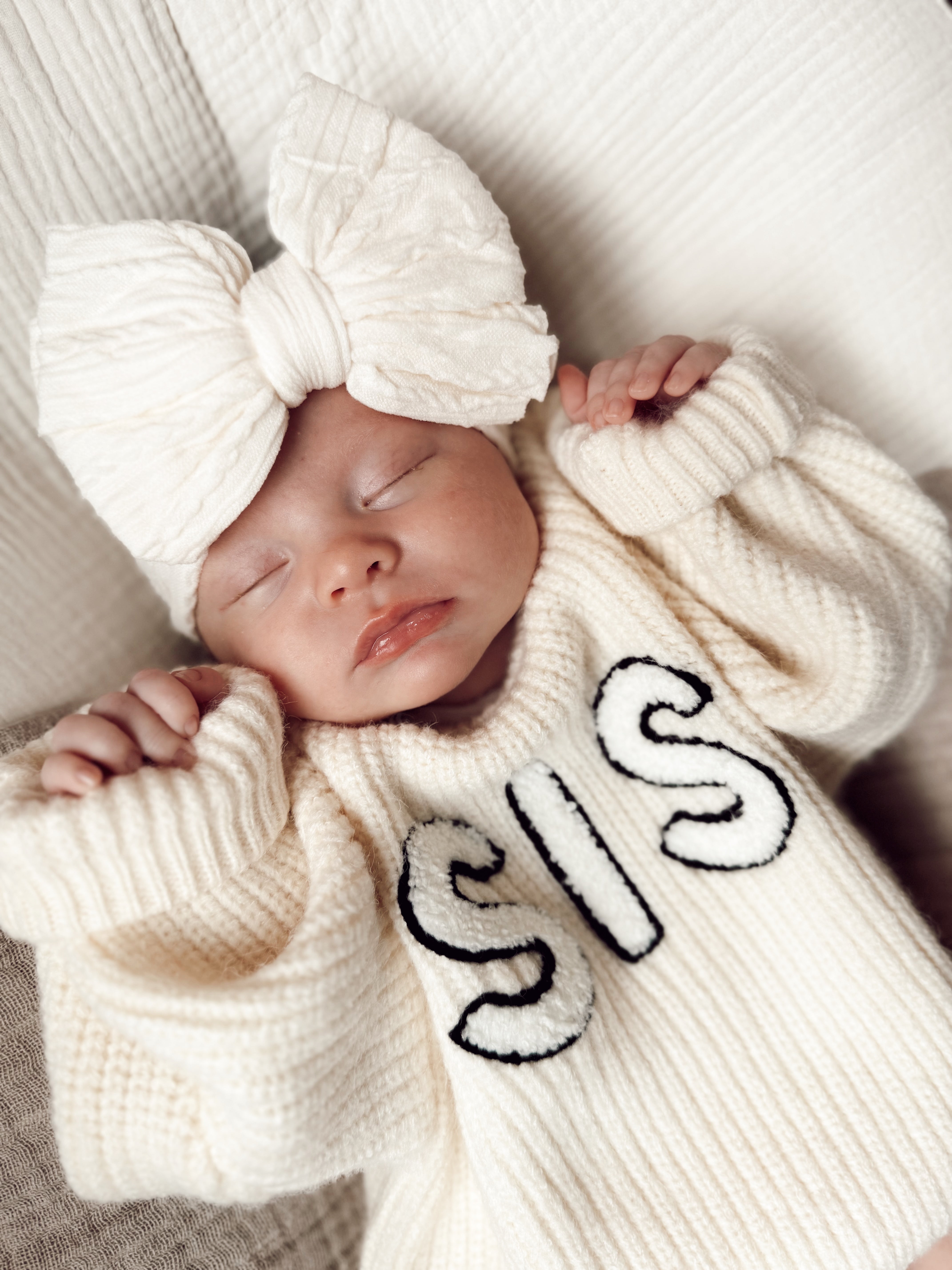 Sleepy baby wearing a cozy cream sweater with "SIS" text and a large bow headband.