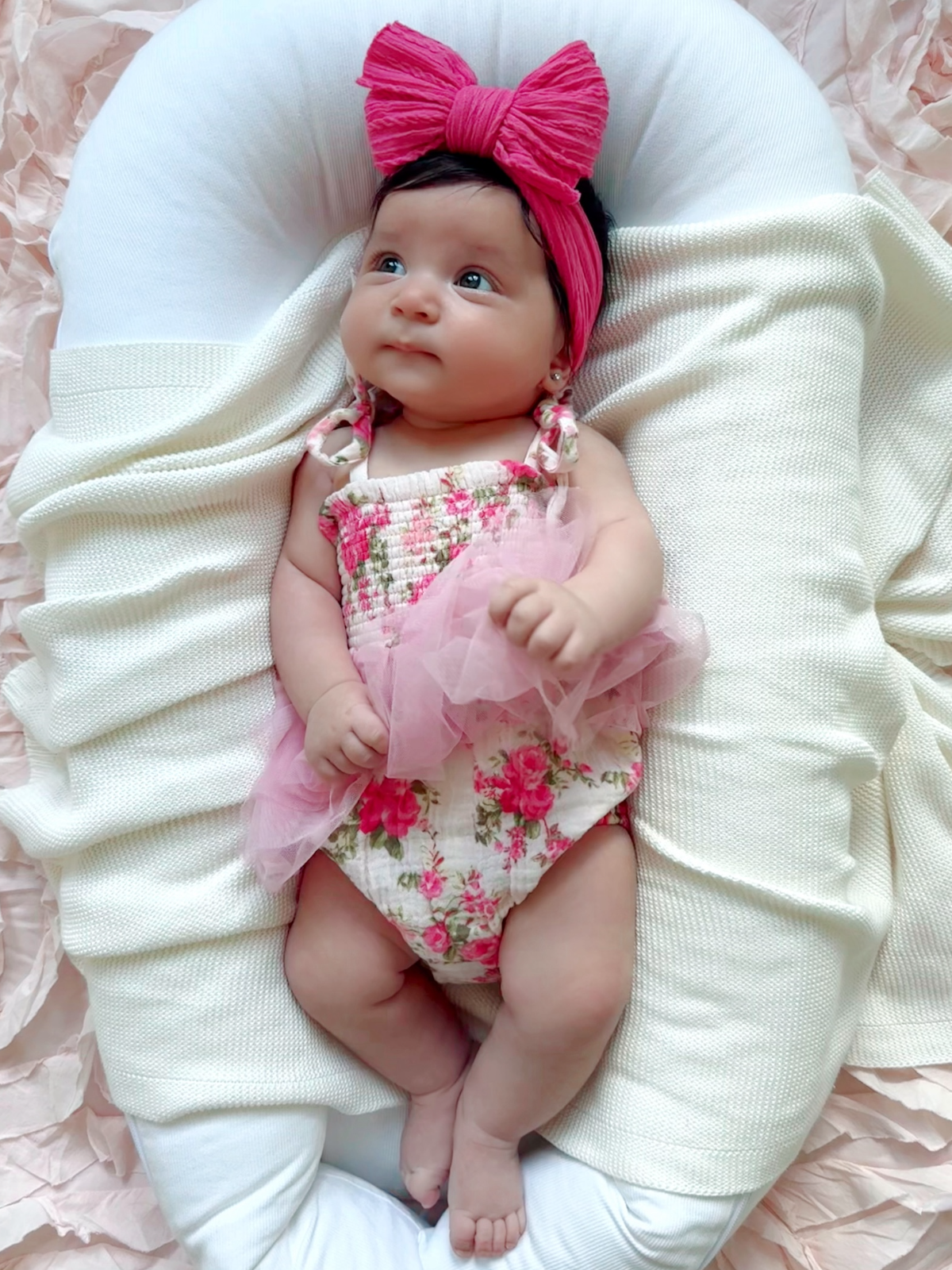 Baby girl in floral outfit and pink bow, lying on a cozy white blanket with soft pink background.