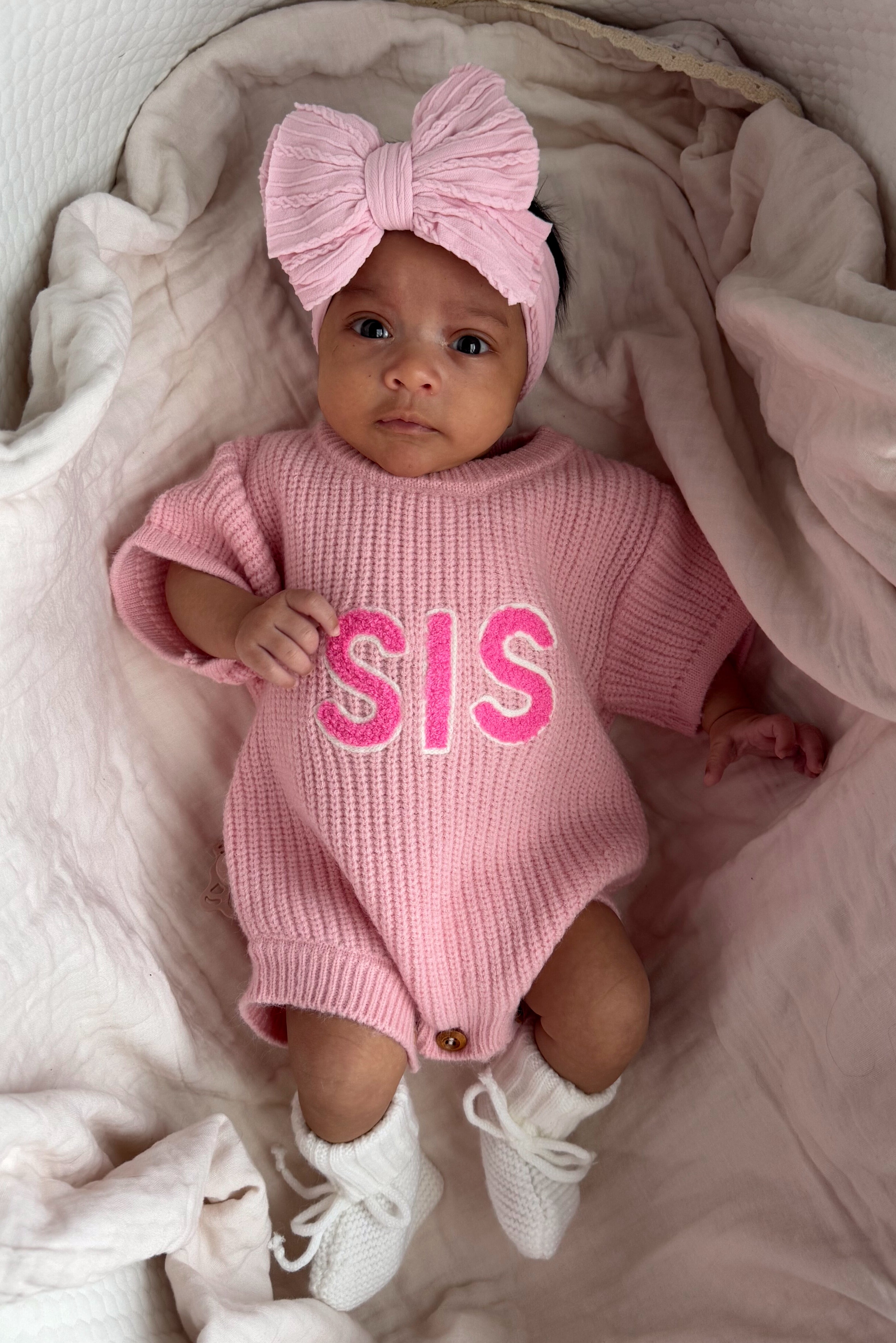 Baby girl in a pink knit outfit with "SIS" text, wearing a matching bow headband and white booties, lying on soft fabric.