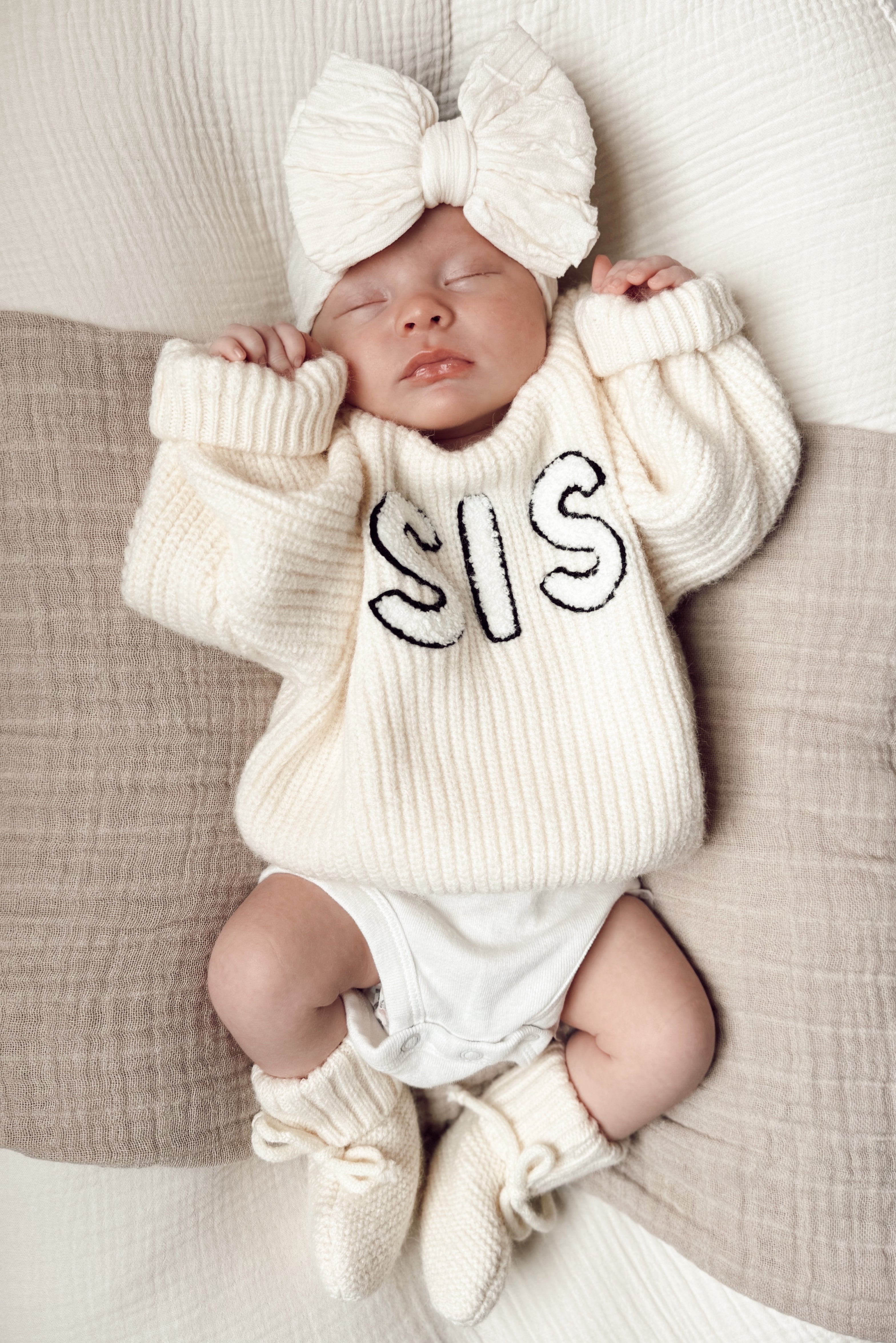 Baby girl in cozy cream sweater with "SIS" print, large bow headband, sleeping on a soft blanket.
