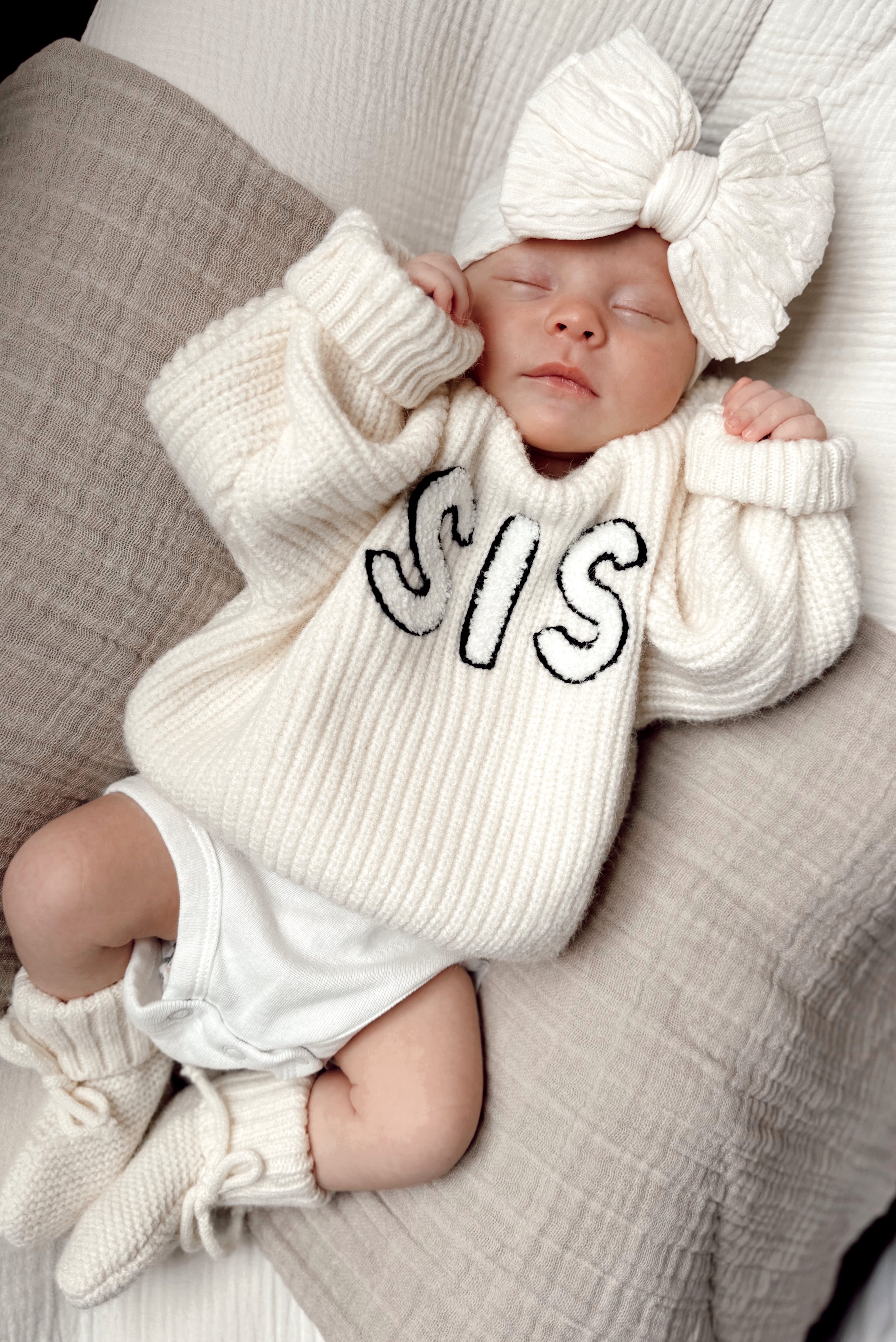 Baby wearing a cozy white sweater with "SIS" printed, resting on soft pillows with a large bow headband.