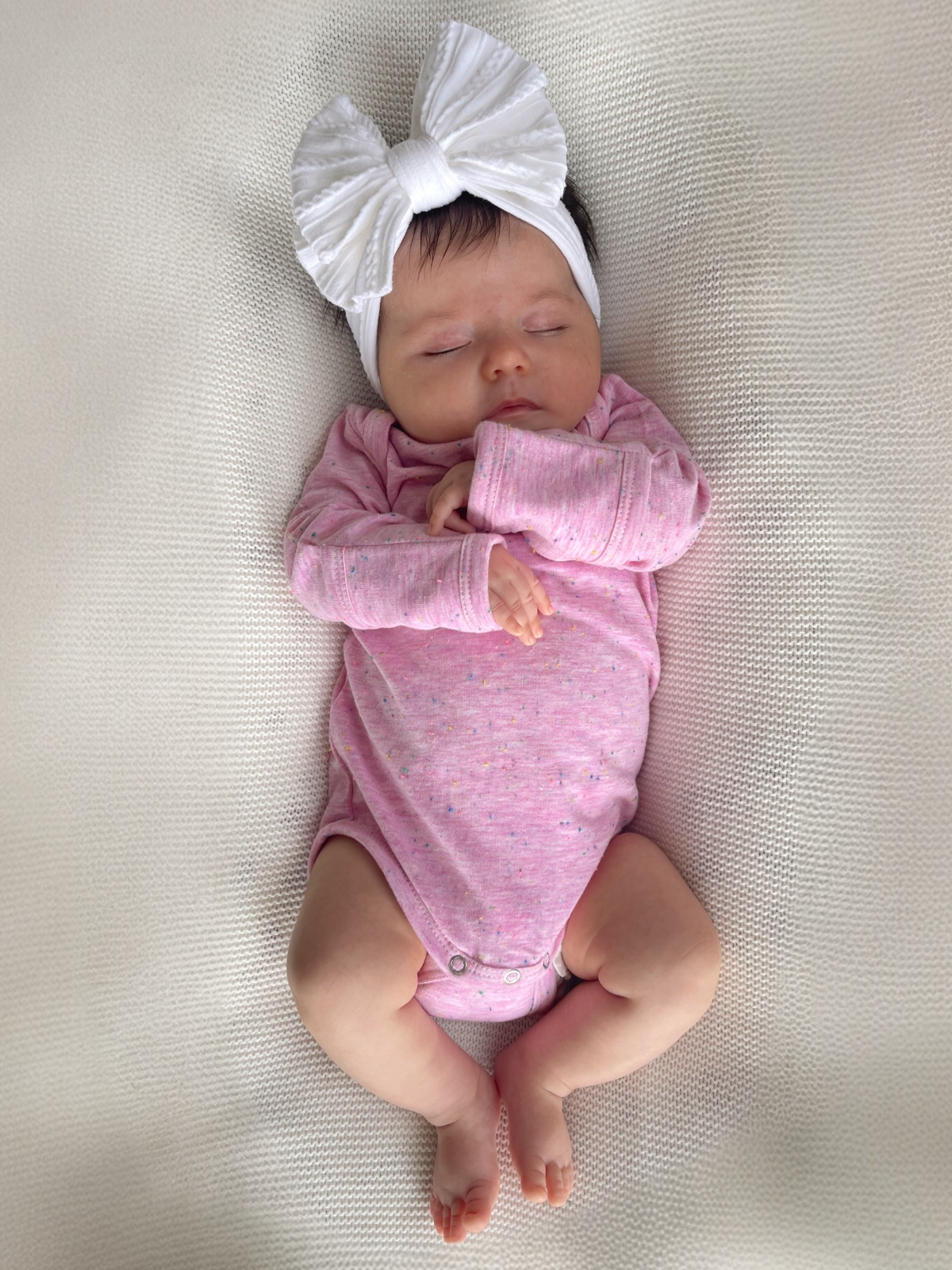 Baby girl in a pink onesie and large white bow headband, sleeping on a textured white blanket.