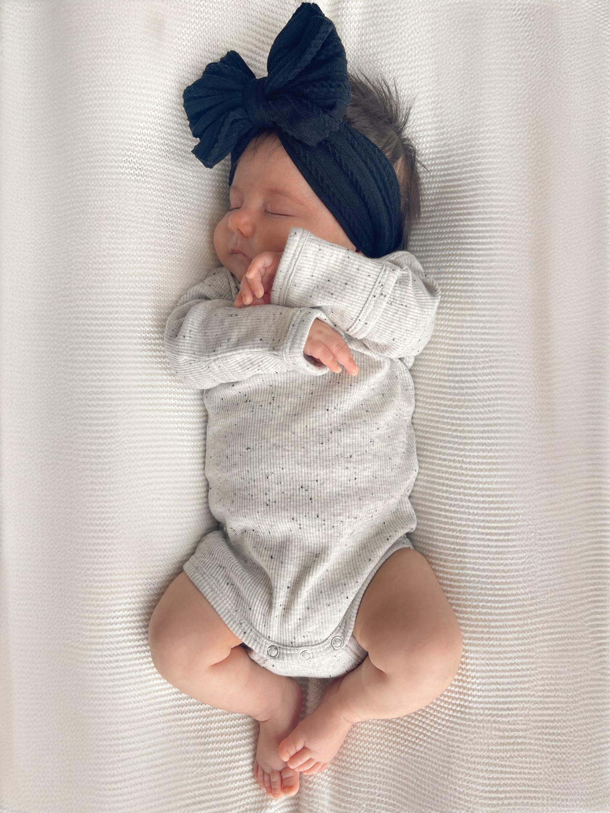 Newborn baby girl in a gray onesie with a large black headband, sleeping on a textured white blanket.