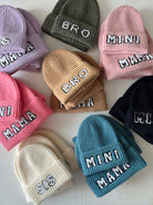 Colorful knit beanies labeled "BRO," "MINI MAMA," "MAMA," "SIS," and more, arranged on a table.