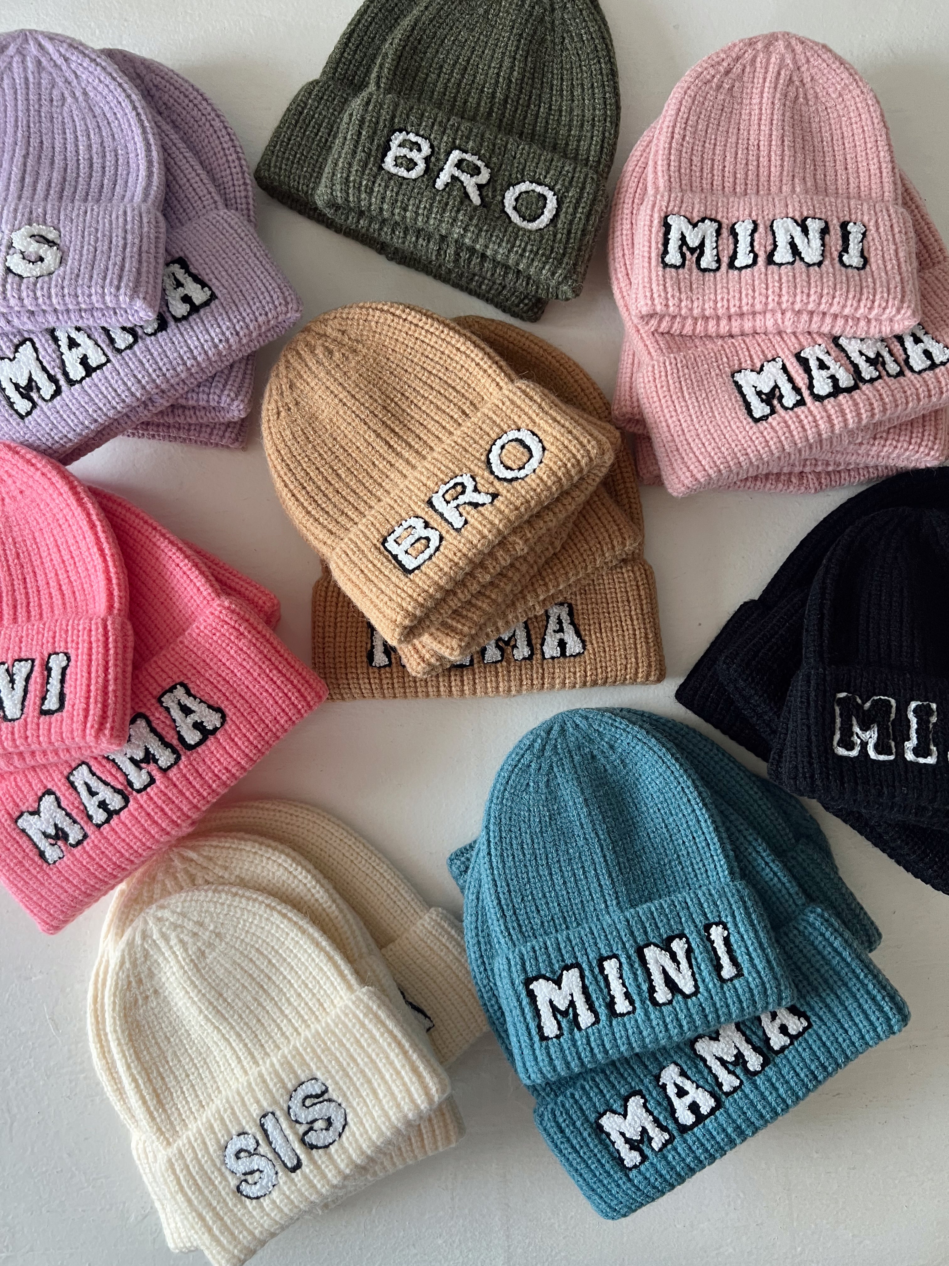 Colorful knit beanies labeled "BRO," "MINI MAMA," "MAMA," "SIS," and more, arranged on a table.
