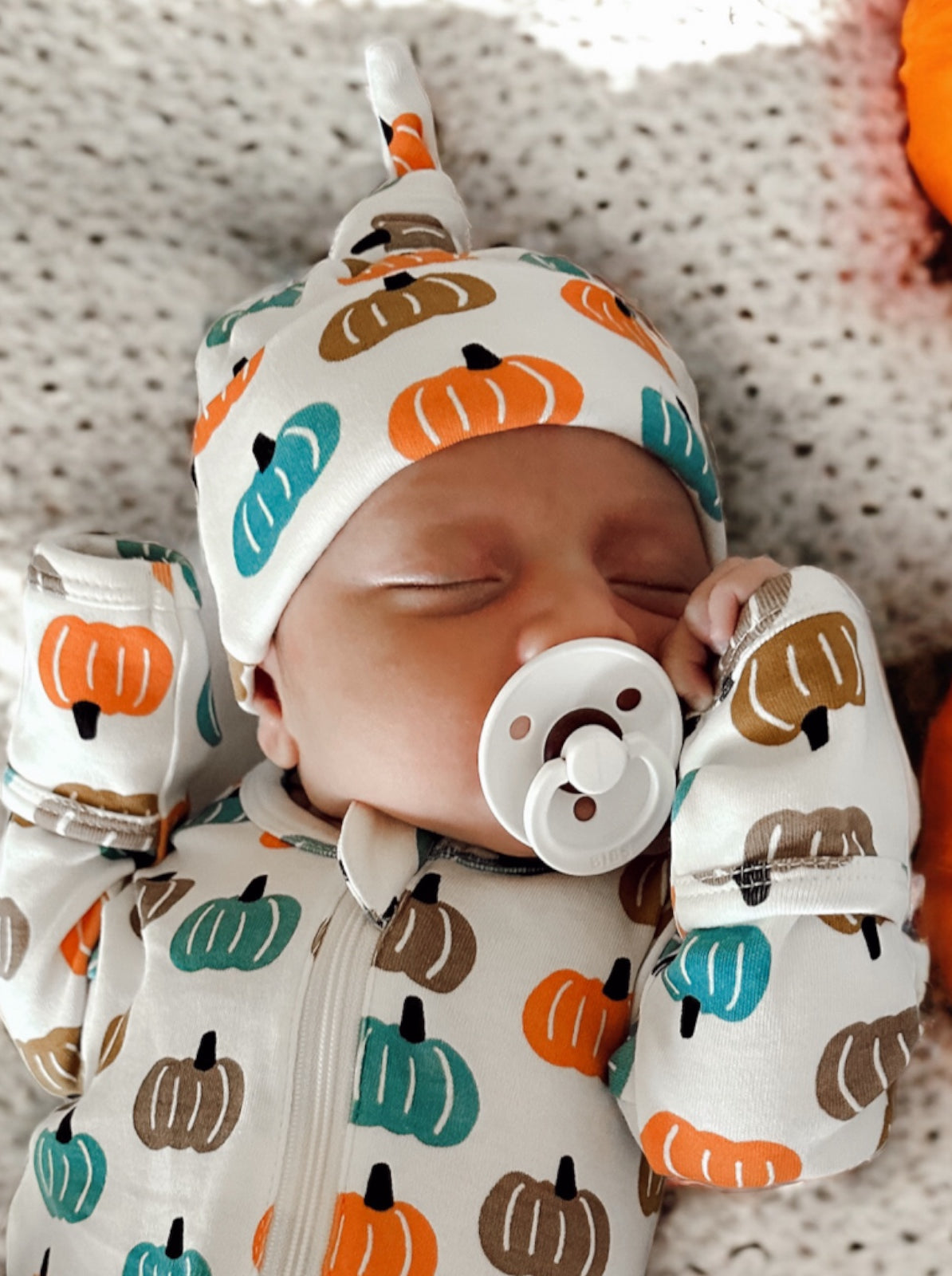 Infant sleeping in a pumpkin-patterned outfit and hat, using a pacifier, on a cozy blanket.