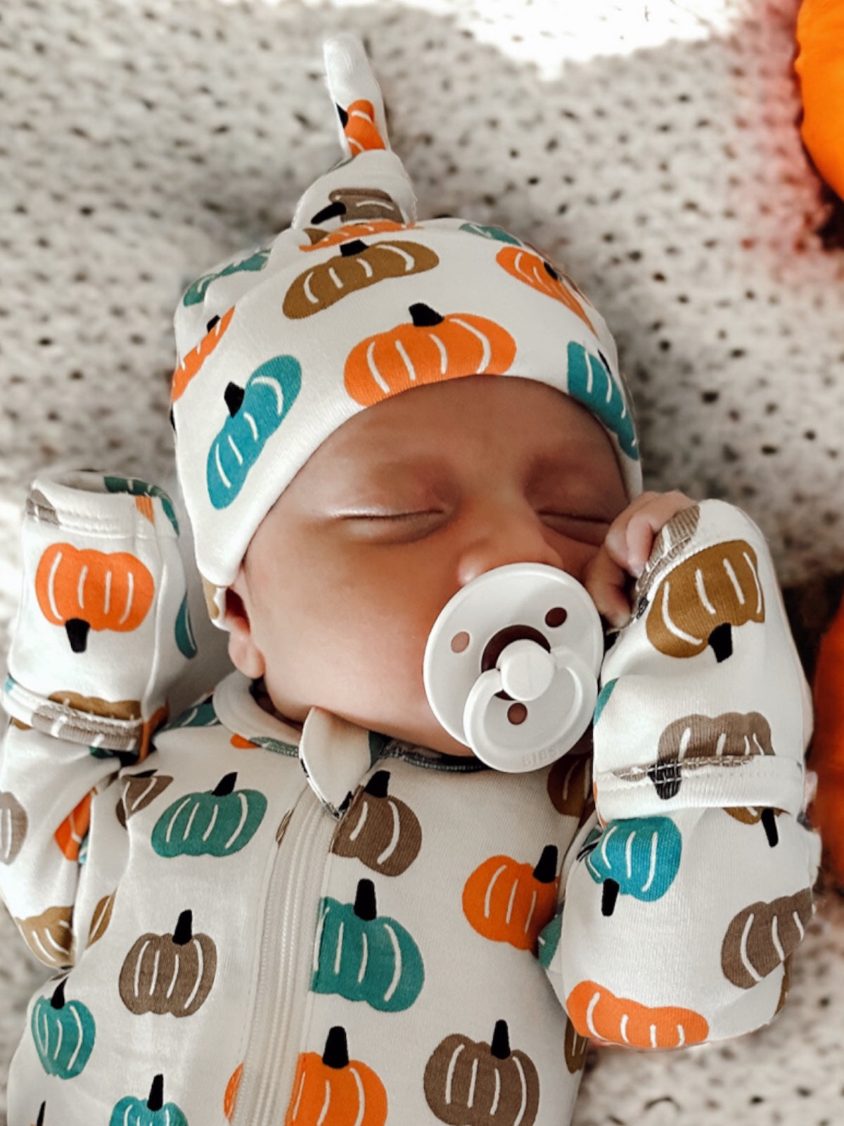 Infant sleeping in a pumpkin-patterned outfit and hat, using a pacifier, on a cozy blanket.