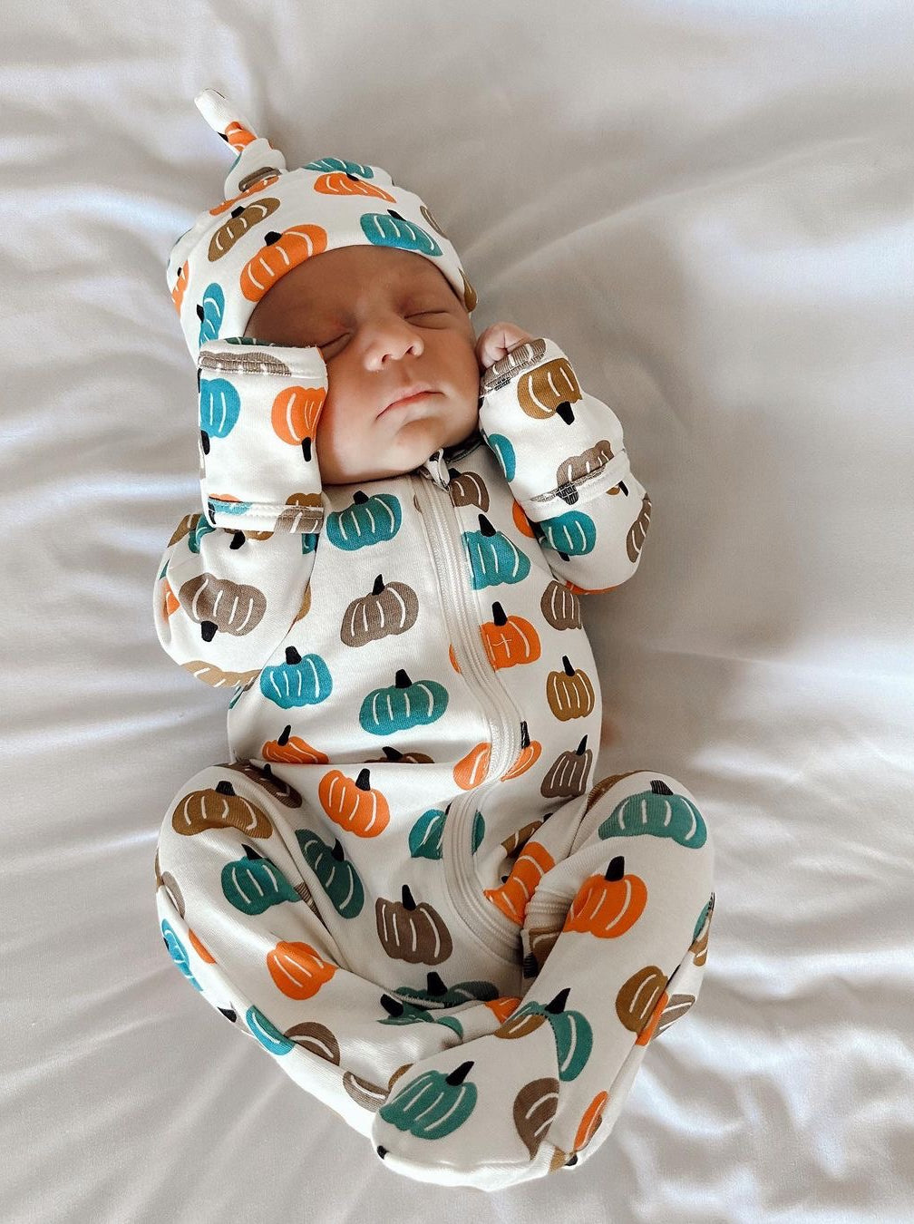 Newborn baby in a pumpkin-patterned onesie and hat, sleeping on a white blanket.