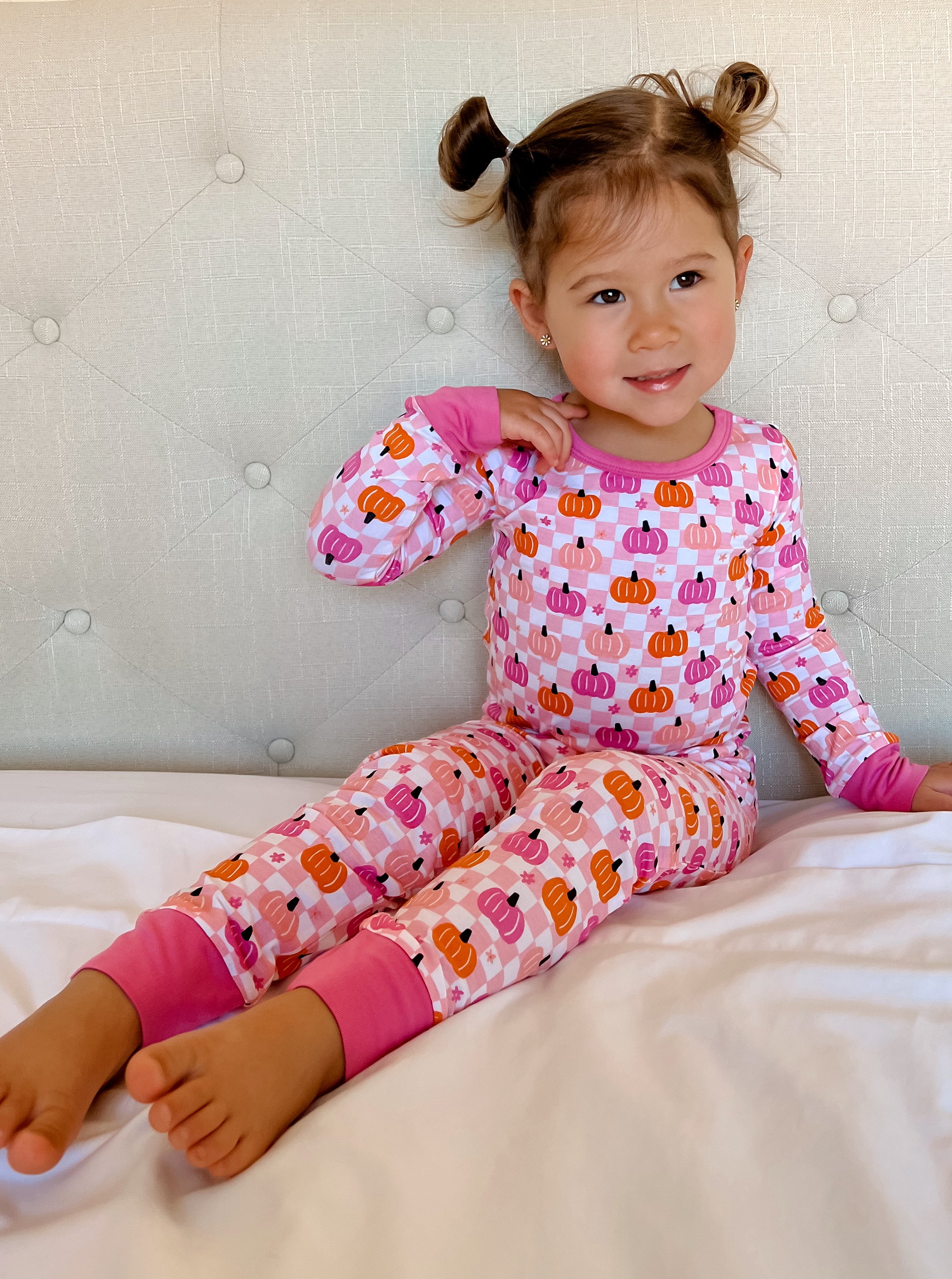Smiling child in pumpkin-patterned pajamas sitting on a bed, with a light gray tufted headboard in the background.