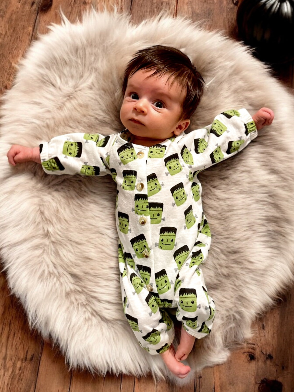 Baby lying on a fluffy rug, wearing a Halloween-themed onesie with cartoon Frankenstein faces.