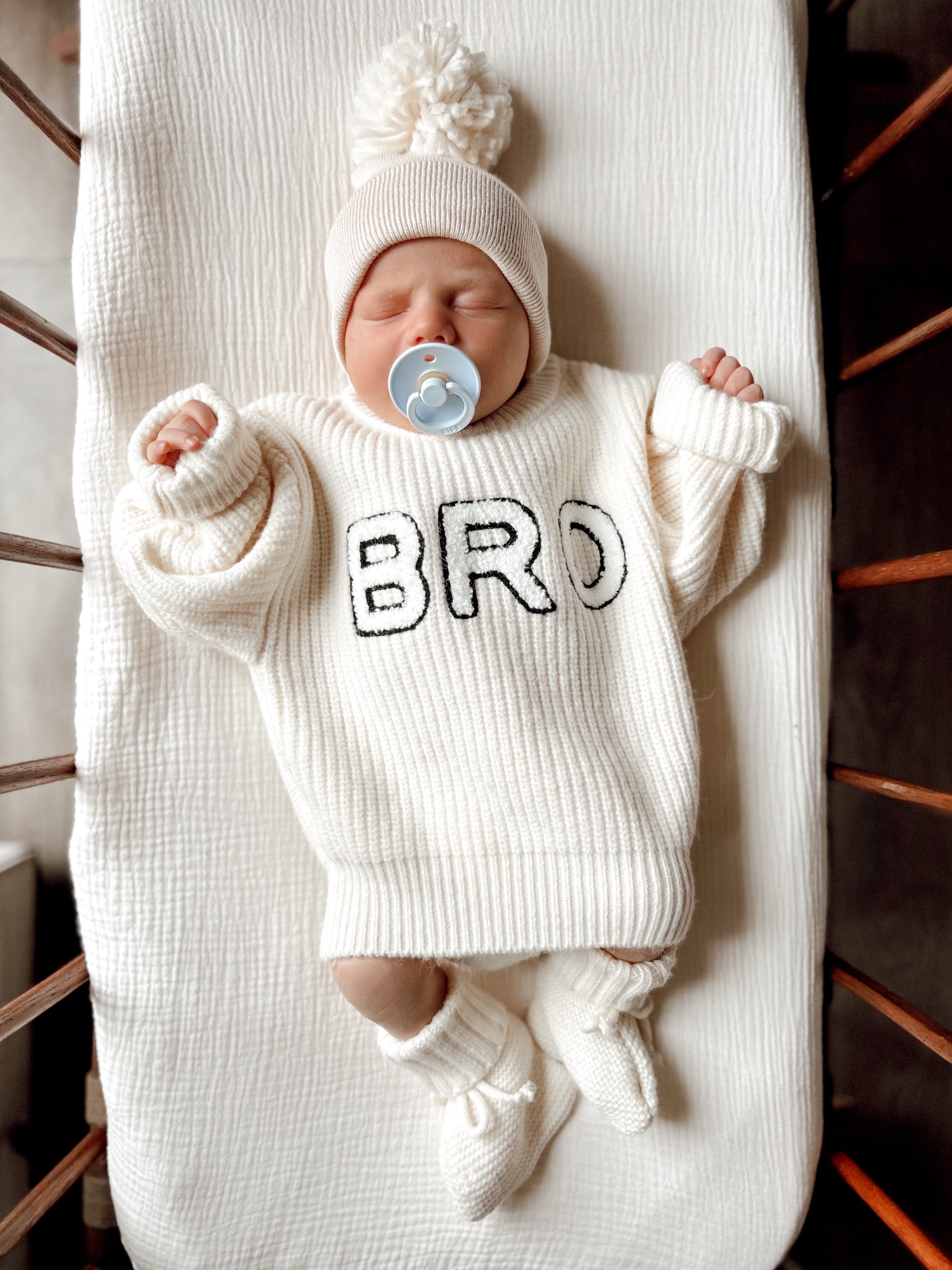 Baby in a cozy, oversized white sweater with "BRO" text, soothing with a pacifier, lying on a textured blanket.
