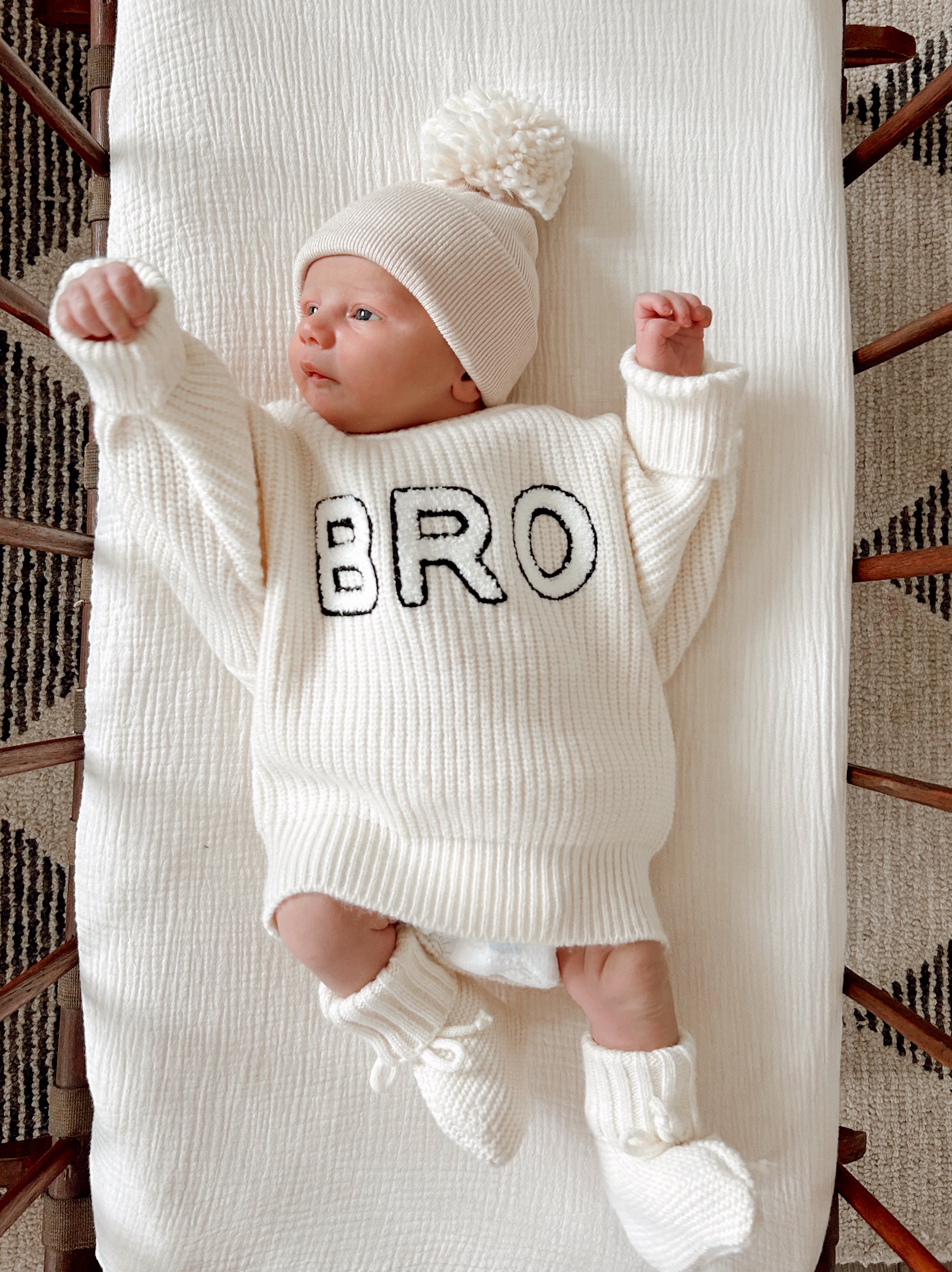 Infant wearing a cream sweater with "BRO" text, a matching beanie, and booties, lying on a textured blanket.