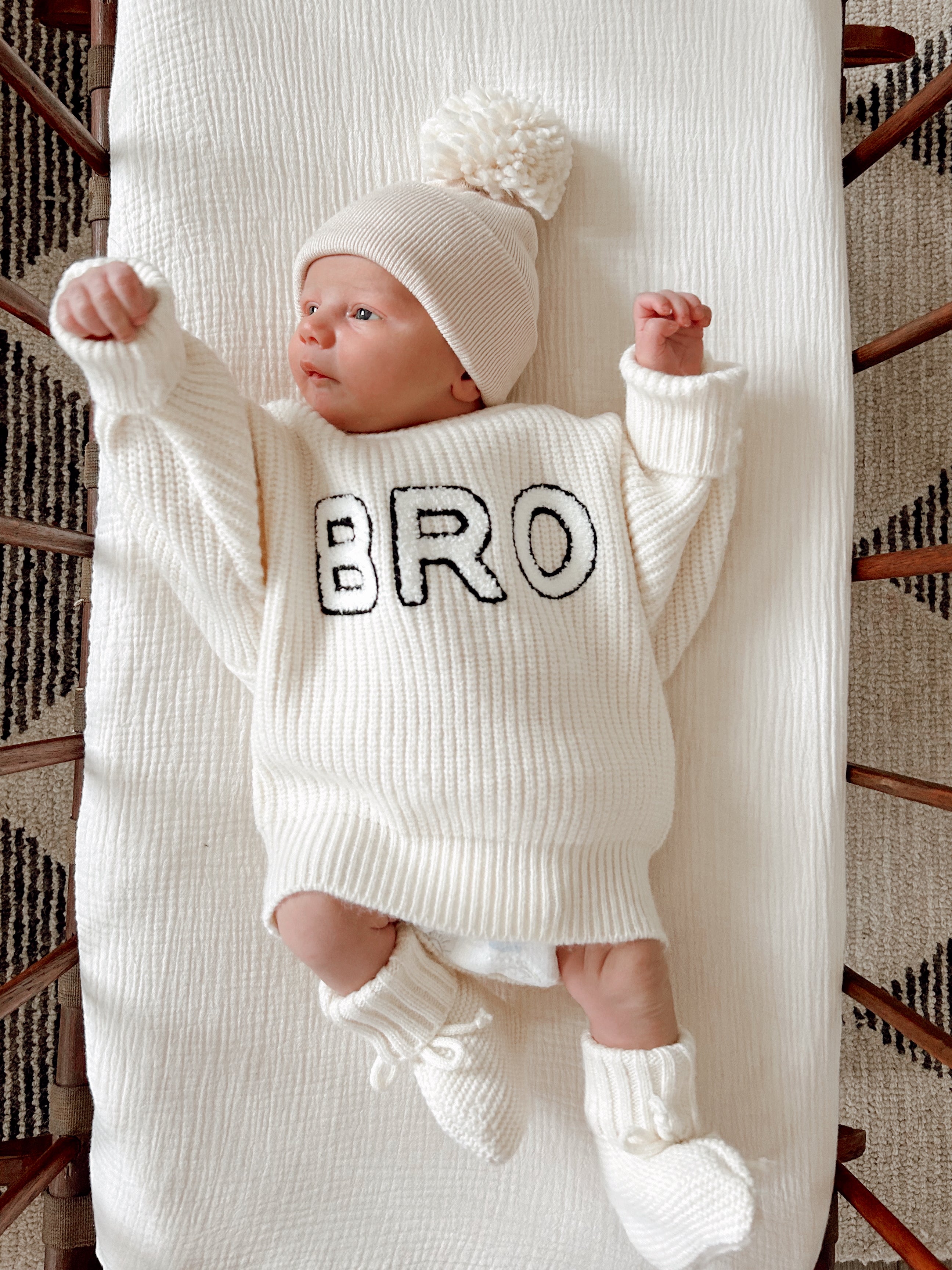 Infant wearing a cream sweater with "BRO" text, a matching beanie, and booties, lying on a textured blanket.