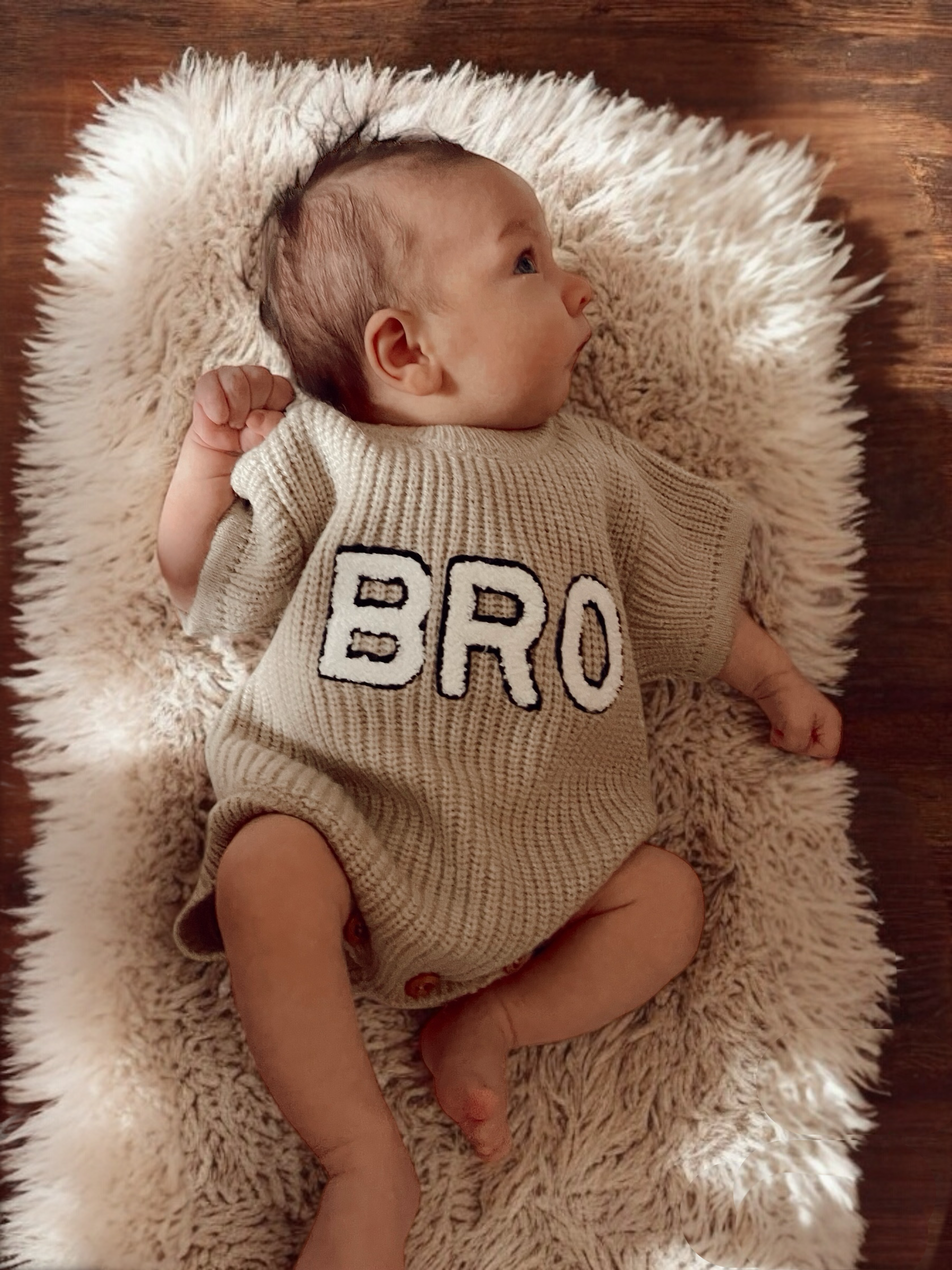 Baby wearing a light tan outfit with "BRO" printed on it, lying on a fluffy rug with a wooden floor background.