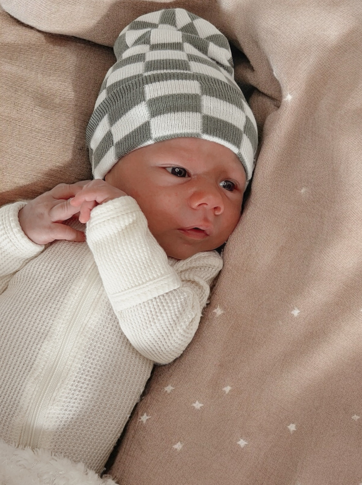 Newborn baby in a white outfit and checkered green hat, resting on a soft blanket with stars.