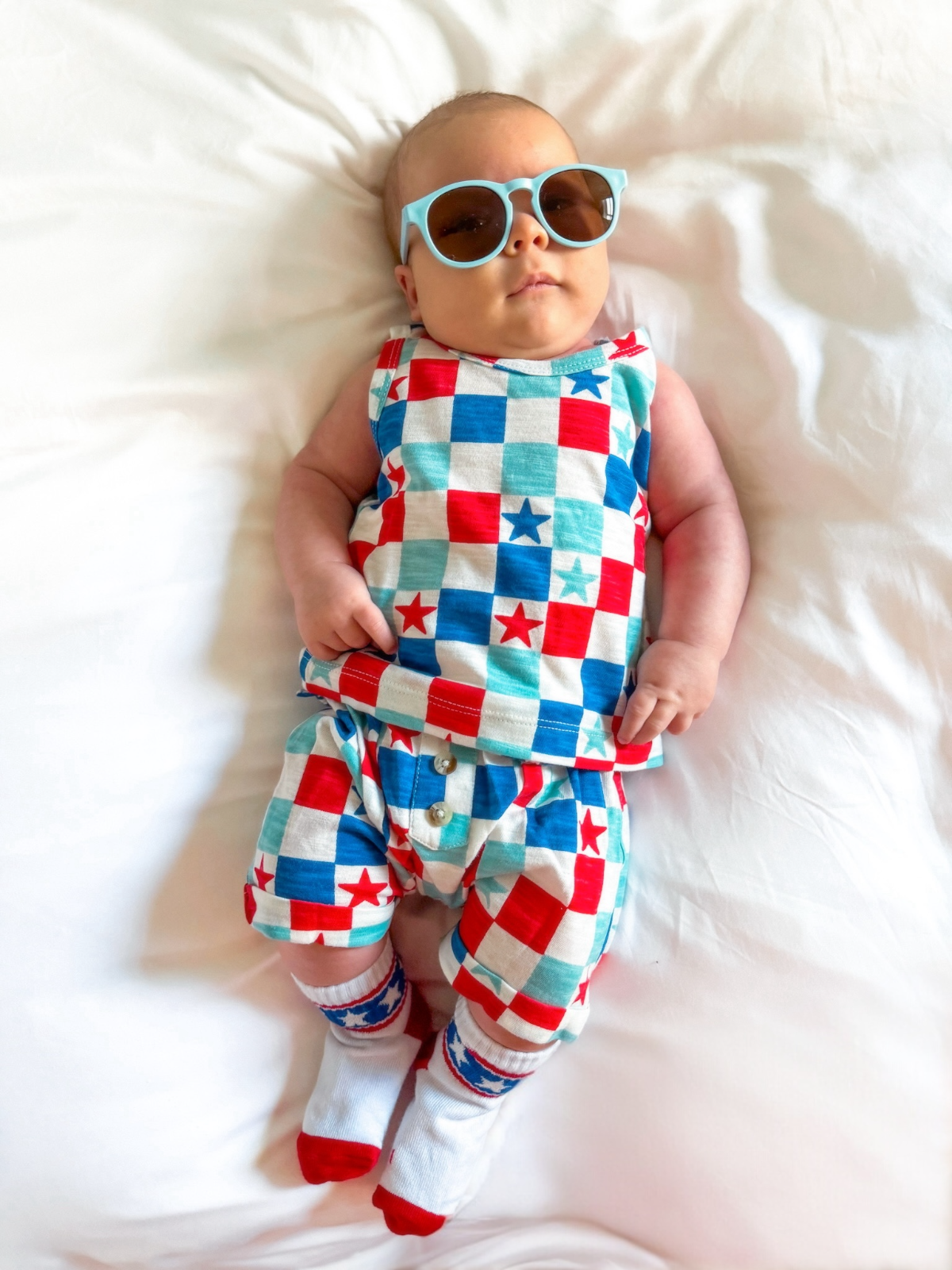 Baby wearing festive outfit with stars and checks, stylish sunglasses, lying on a white bedspread.