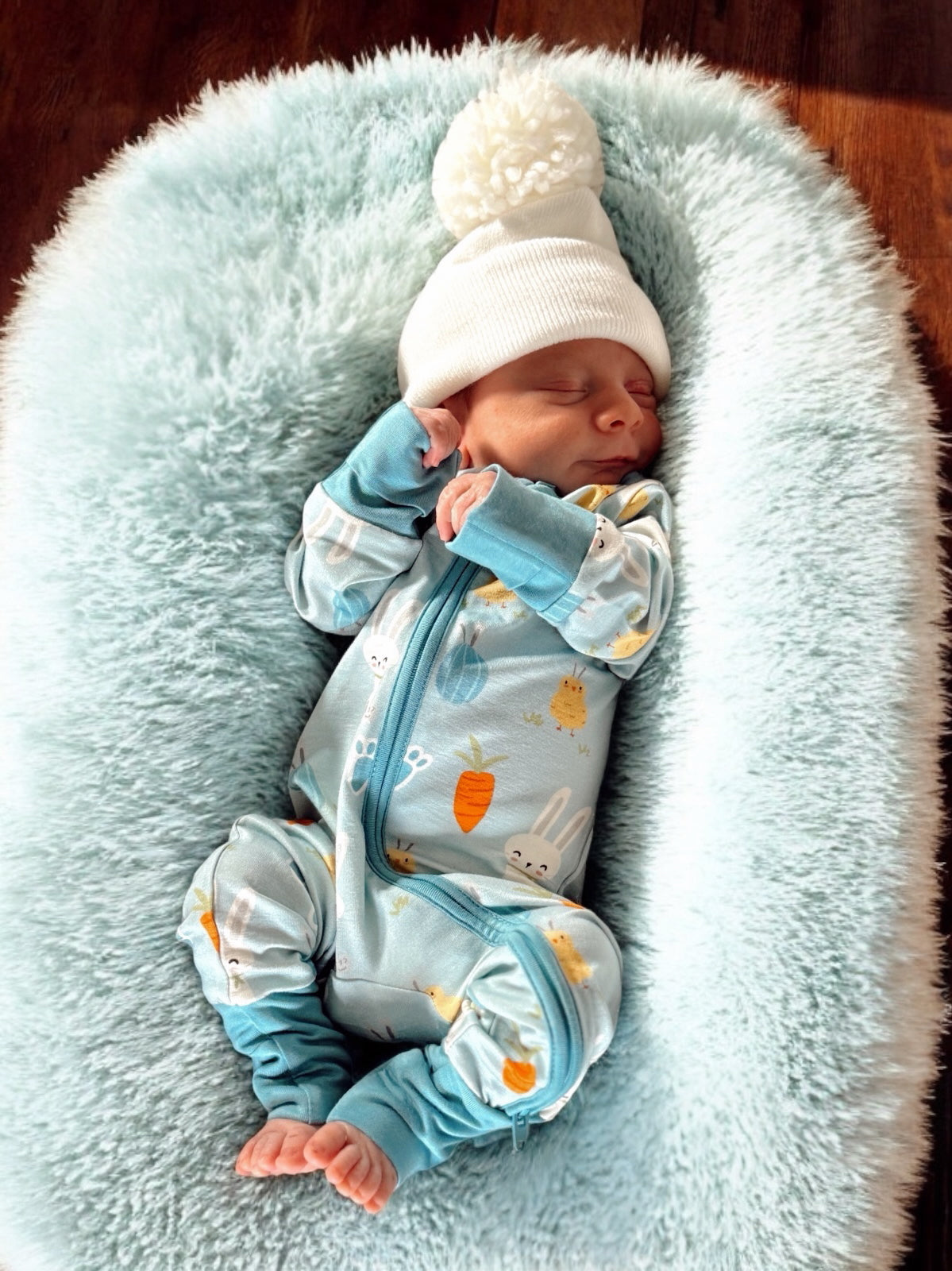 Sleeping newborn swaddled in a cozy blanket wearing a bunny-themed onesie and a white pom-pom beanie.