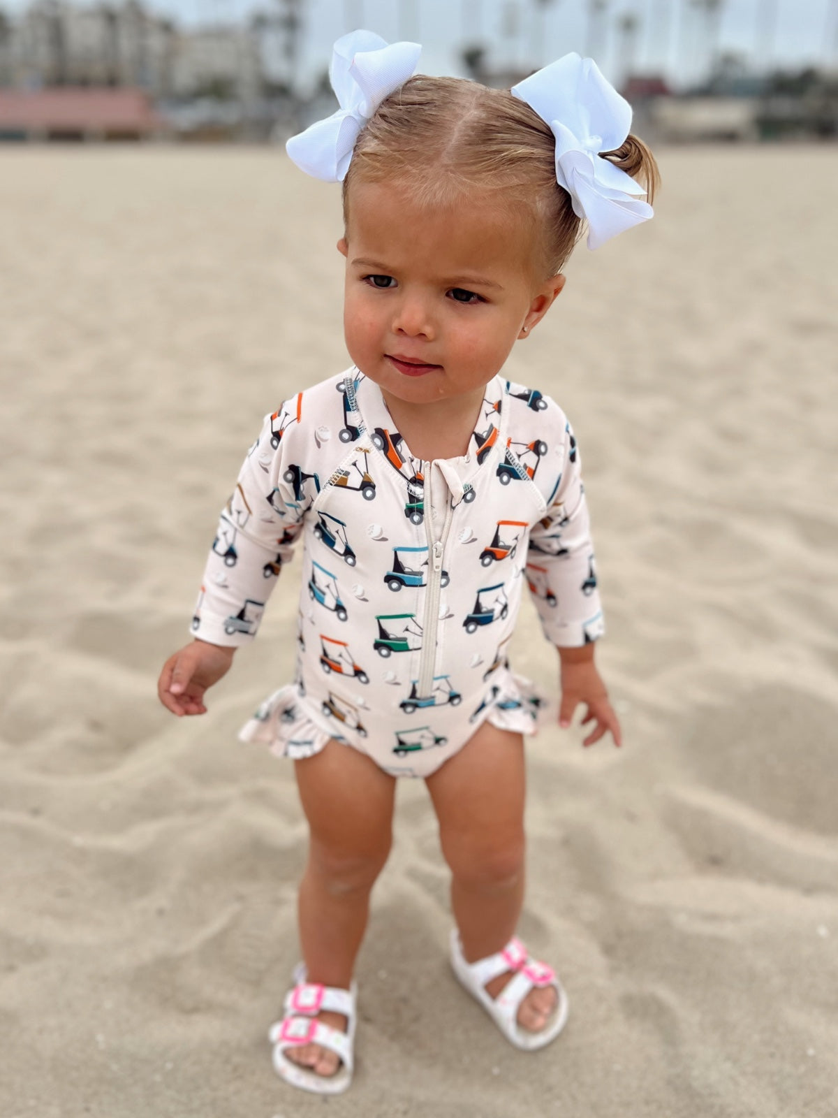 Toddler in a beach outfit with golf cart pattern, wearing a big bow, standing on sandy beach.