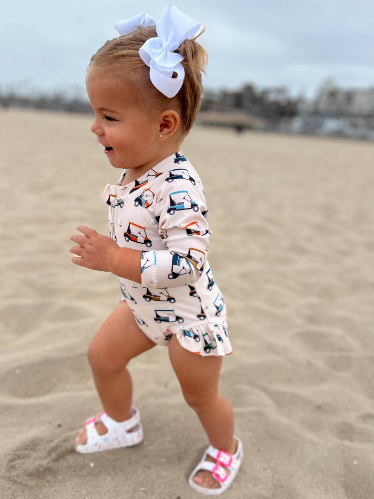 Toddler in playful car-patterned outfit and white bow, joyfully running on sandy beach.