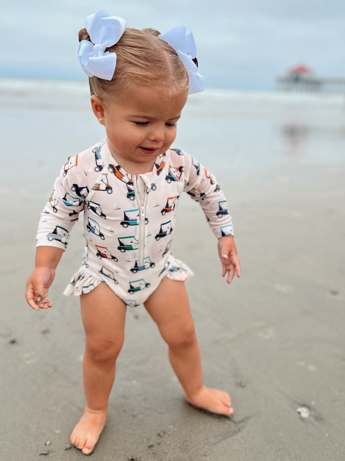 Toddler wading in the sand at the beach, wearing a colorful swimsuit and large bows in her hair.