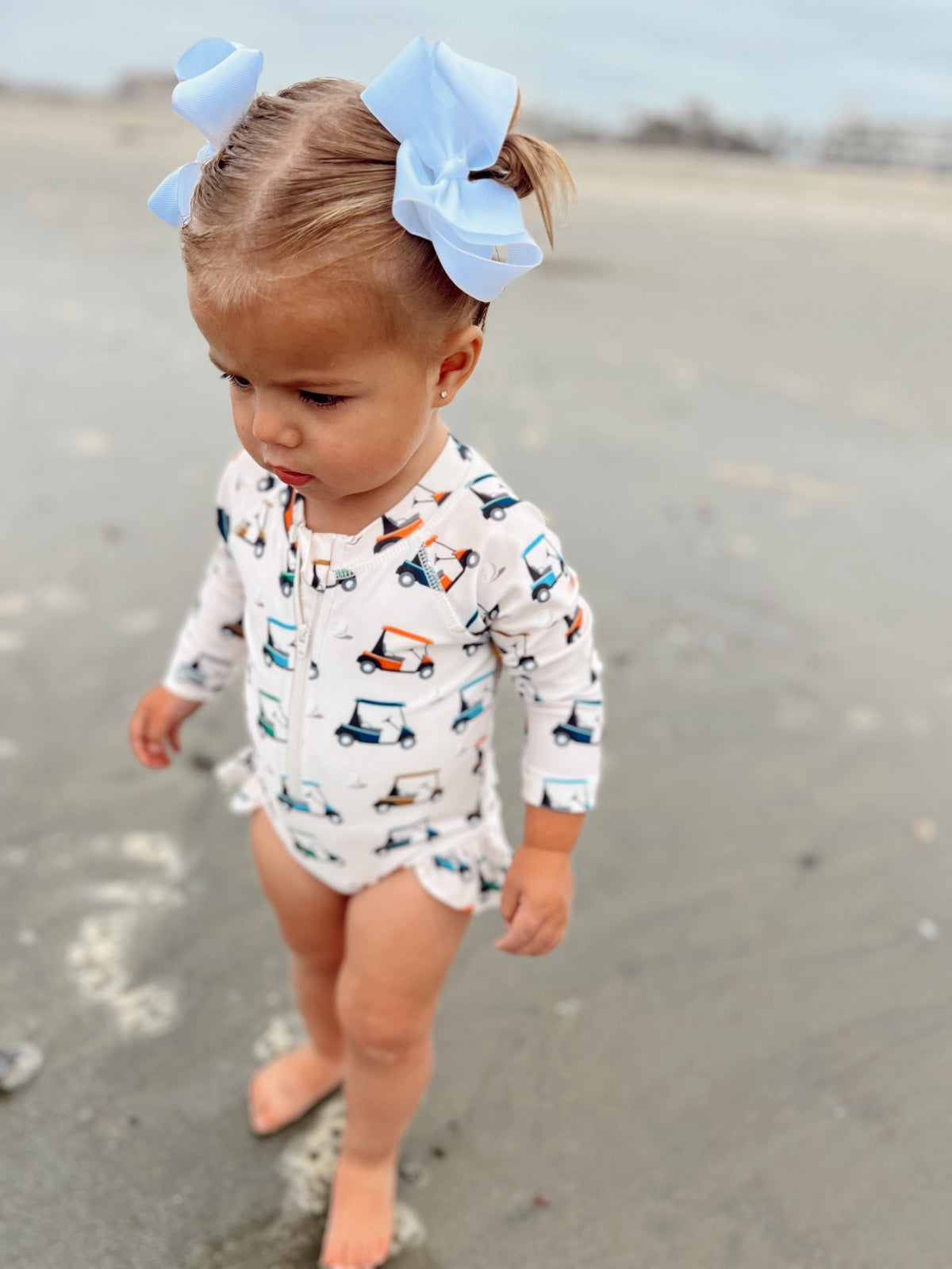 Toddler on the beach wearing a printed swimsuit and blue hair bows, looking down at the wet sand.