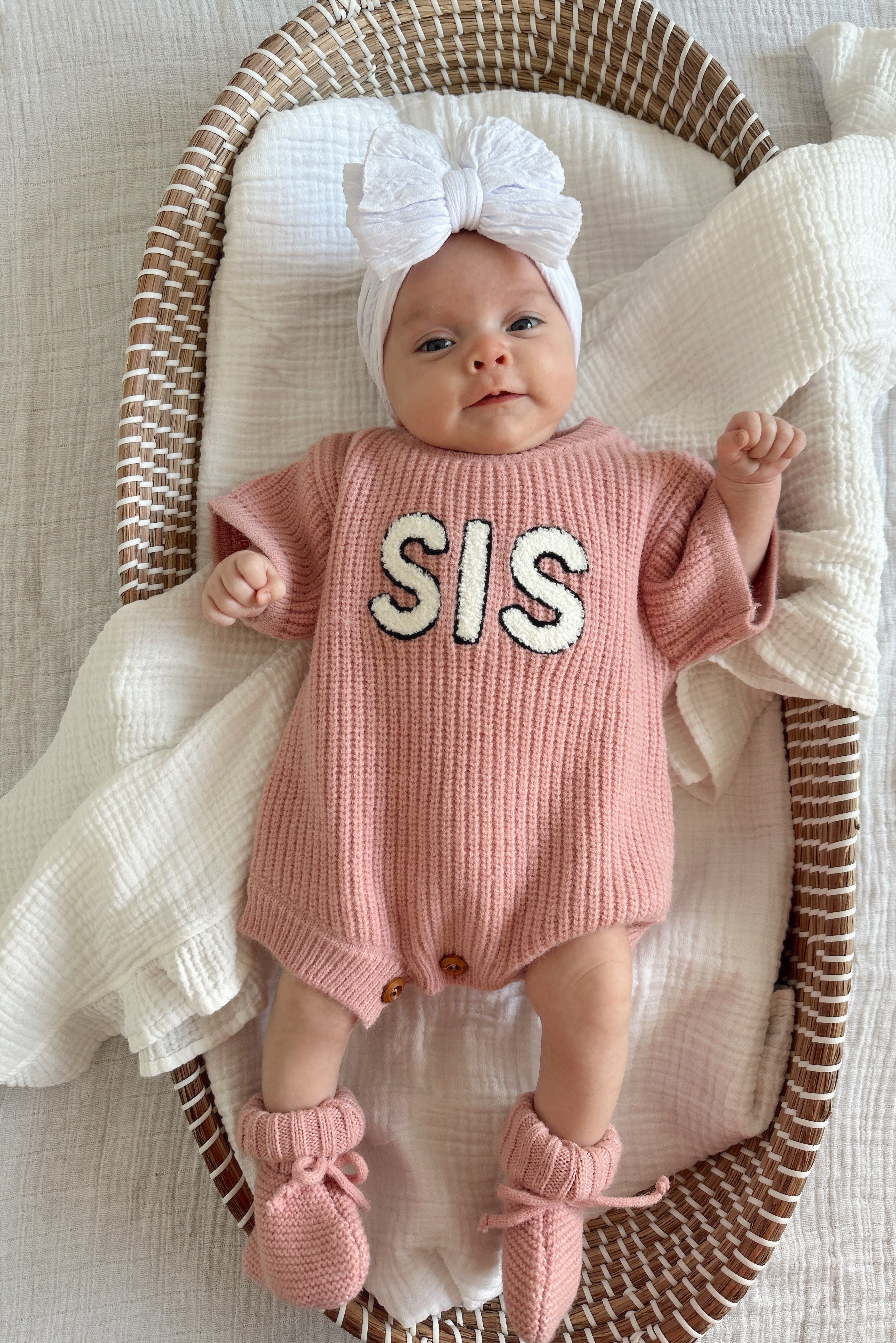 Baby in pink knit romper with "SIS" text, wearing a large white bow and booties, lying in a woven basket.