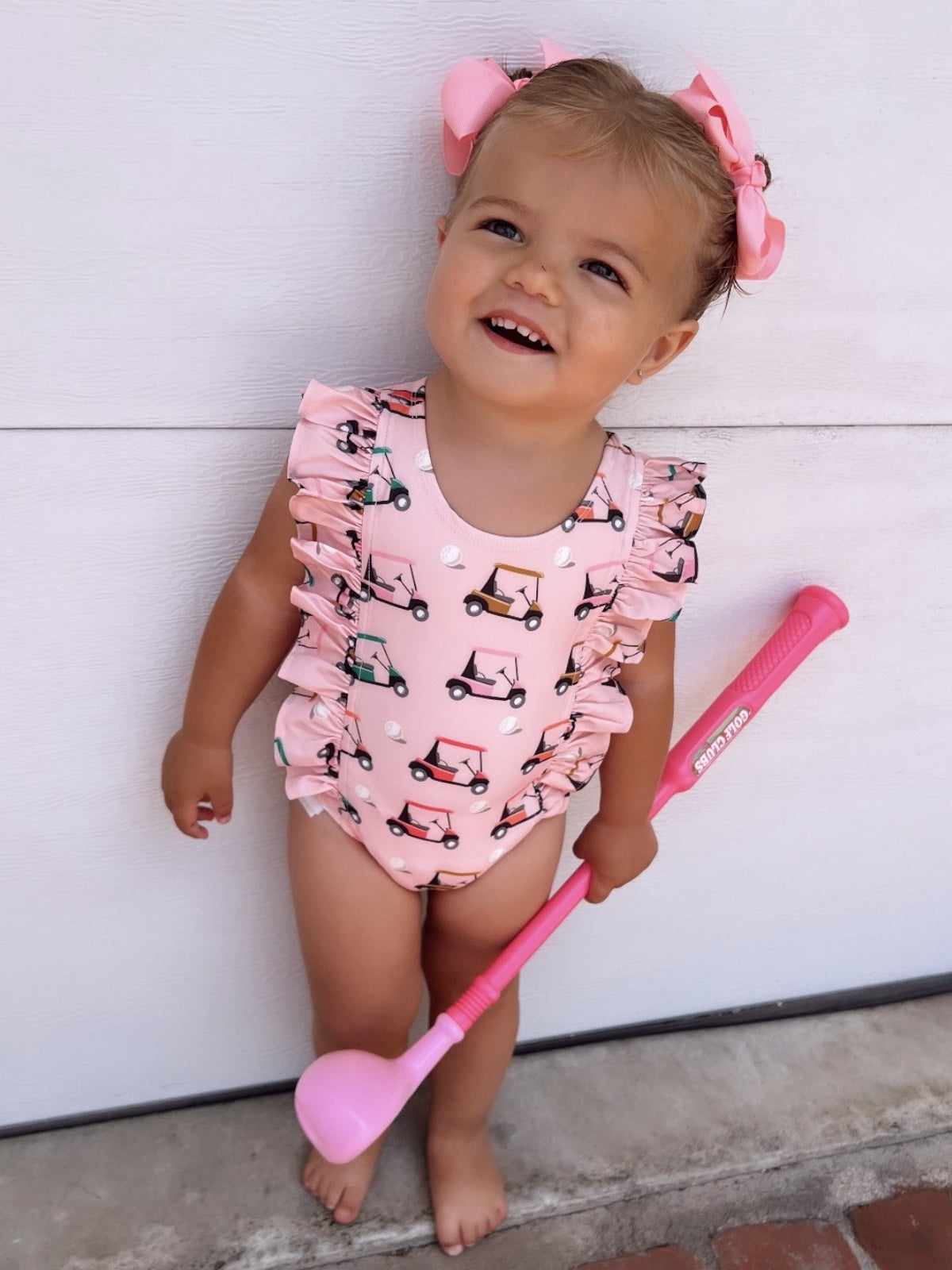 Smiling toddler in a pink golf cart patterned swimsuit, holding a pink golf club, with matching hair bows.