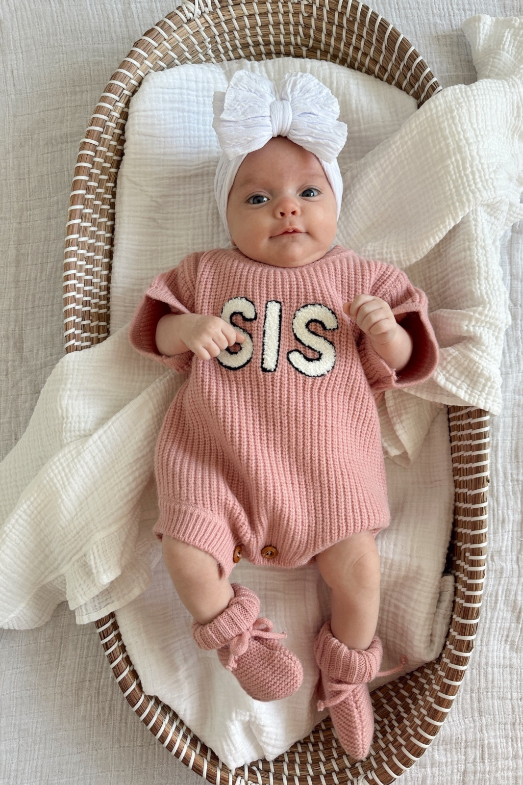 Baby in a cozy pink outfit with "SIS" lettering, nestled in a woven basket with a white blanket.