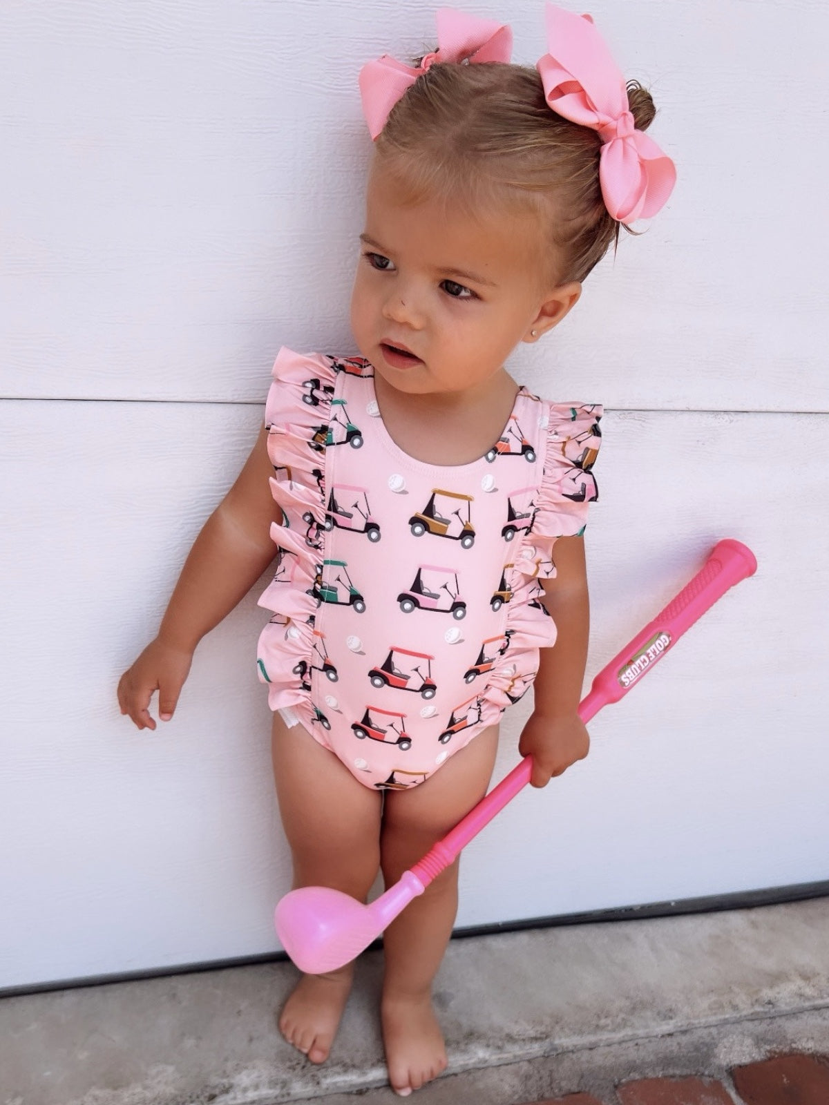 Toddler girl in a pink ruffled swimsuit holding a pink golf club, standing against a white background.
