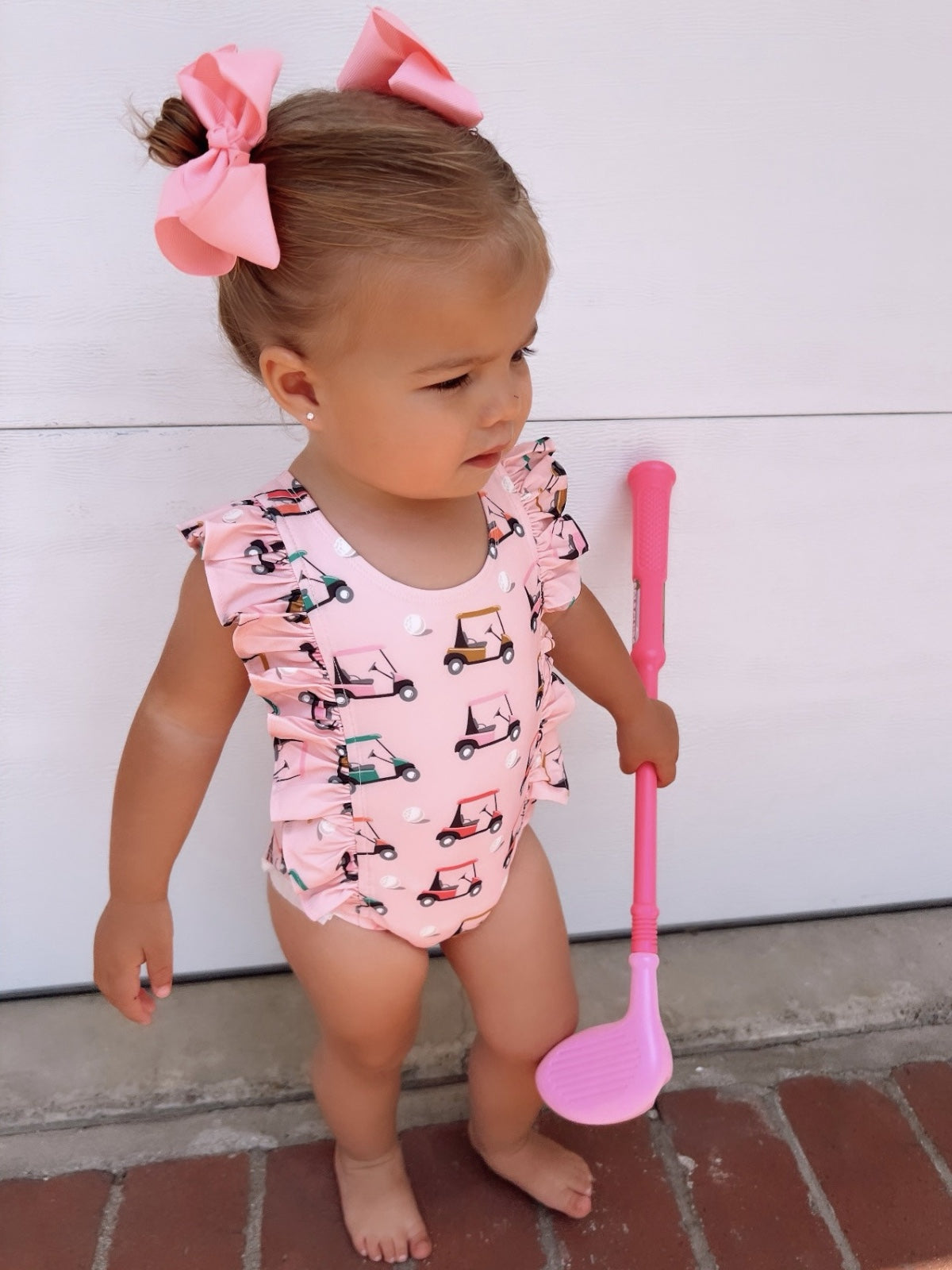 Toddler with pink bow and golf-themed swimsuit, holding a pink golf club, standing on brick pavement.