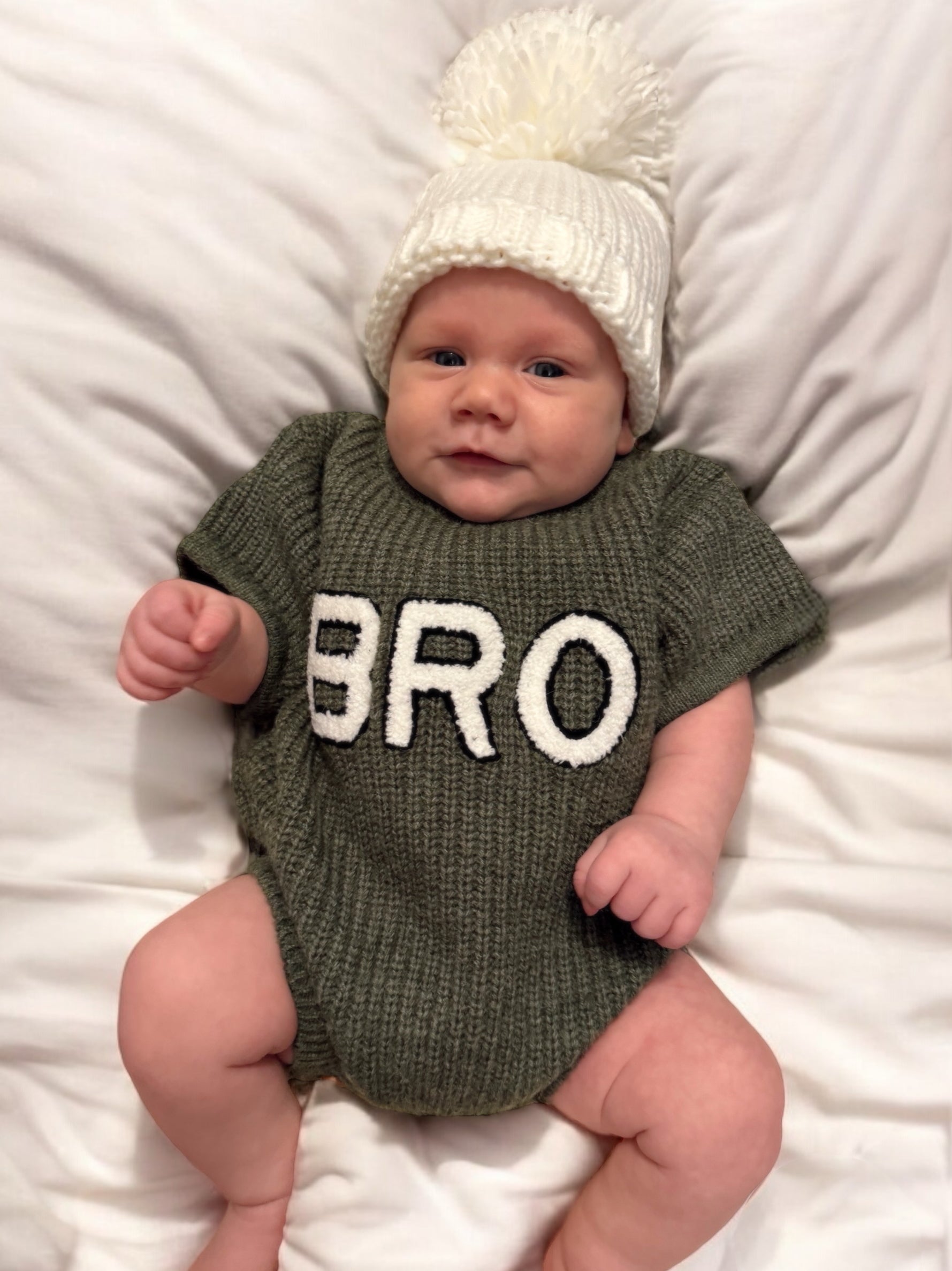 Baby wearing a green sweater with "BRO" printed on it, posing on a white blanket, topped with a white pom-pom hat.