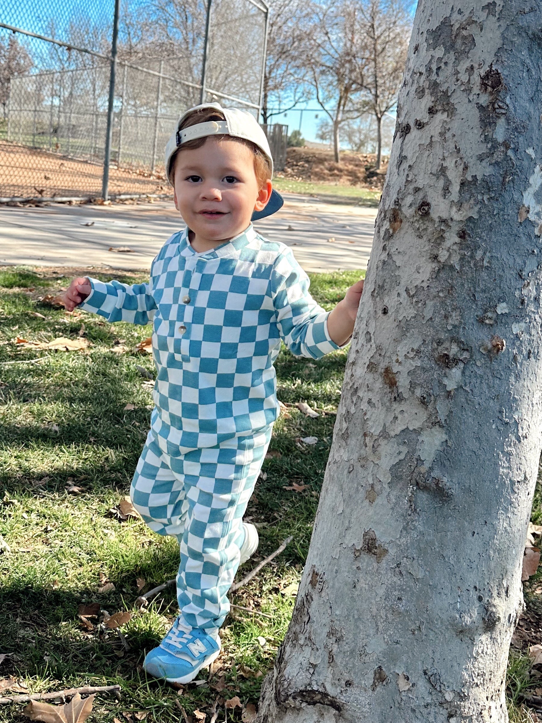 Toddler in a blue and white checkered outfit smiles while standing against a tree in a park.