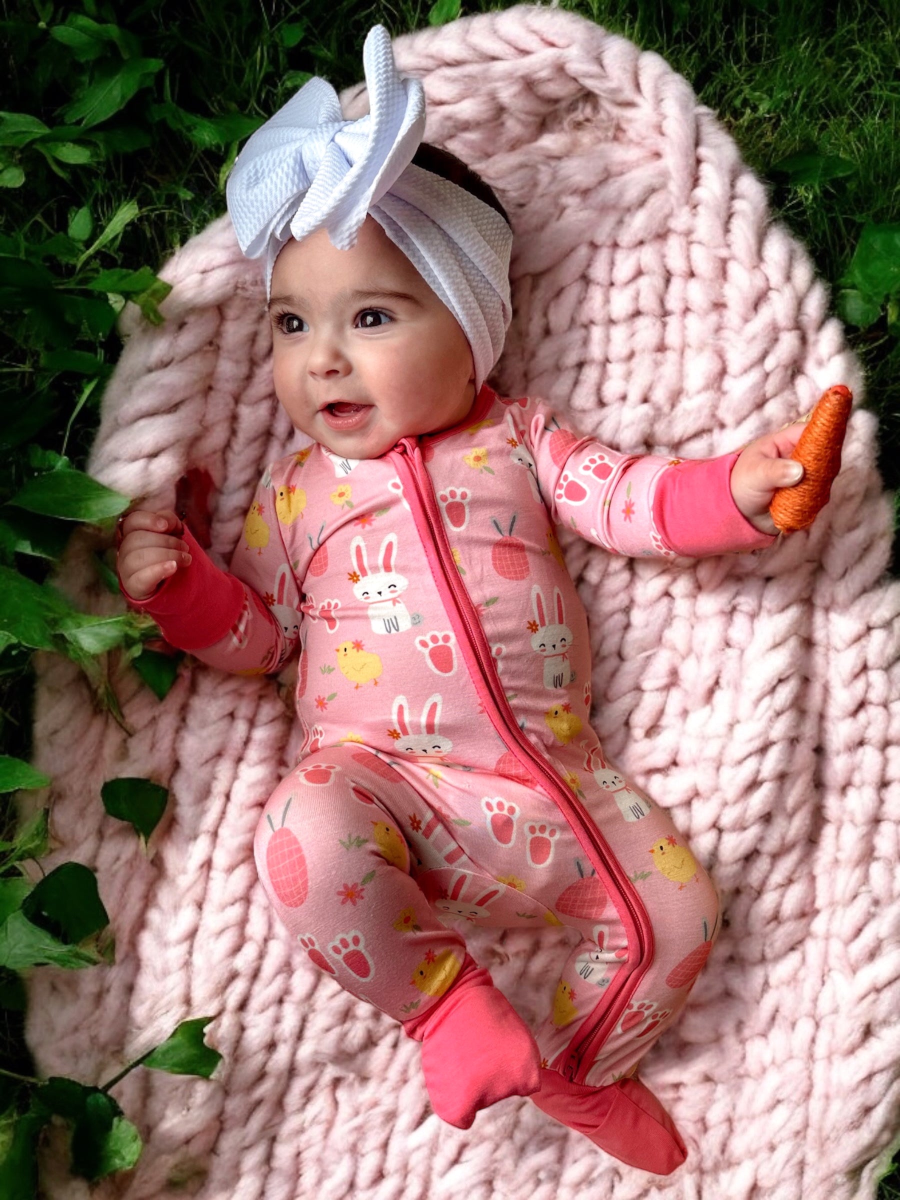 Smiling baby in a pink bunny onesie, holding a carrot on a soft blanket surrounded by greenery.