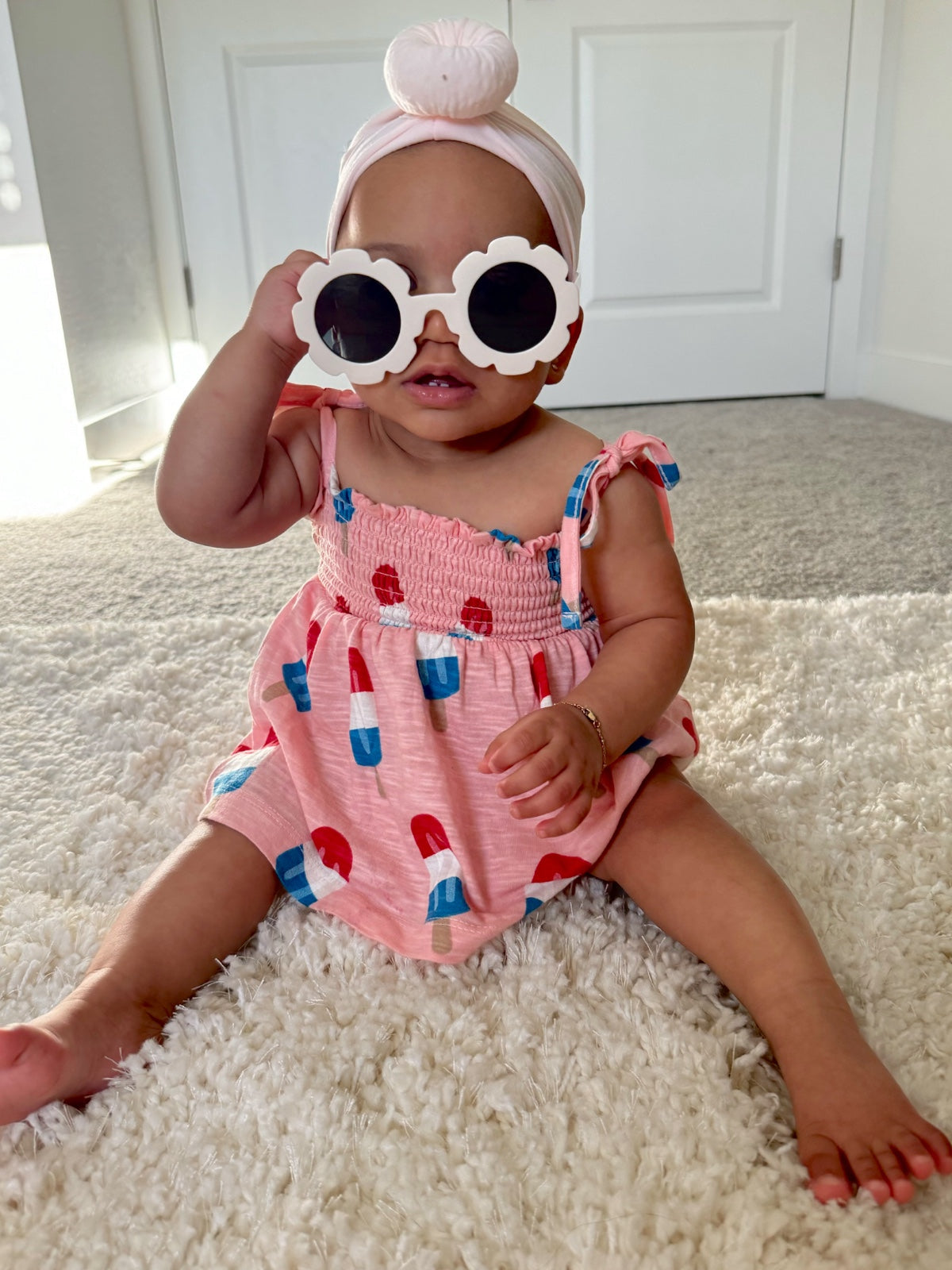 Baby wearing a pink ice cream-patterned outfit and flower-shaped sunglasses, sitting on a soft rug.