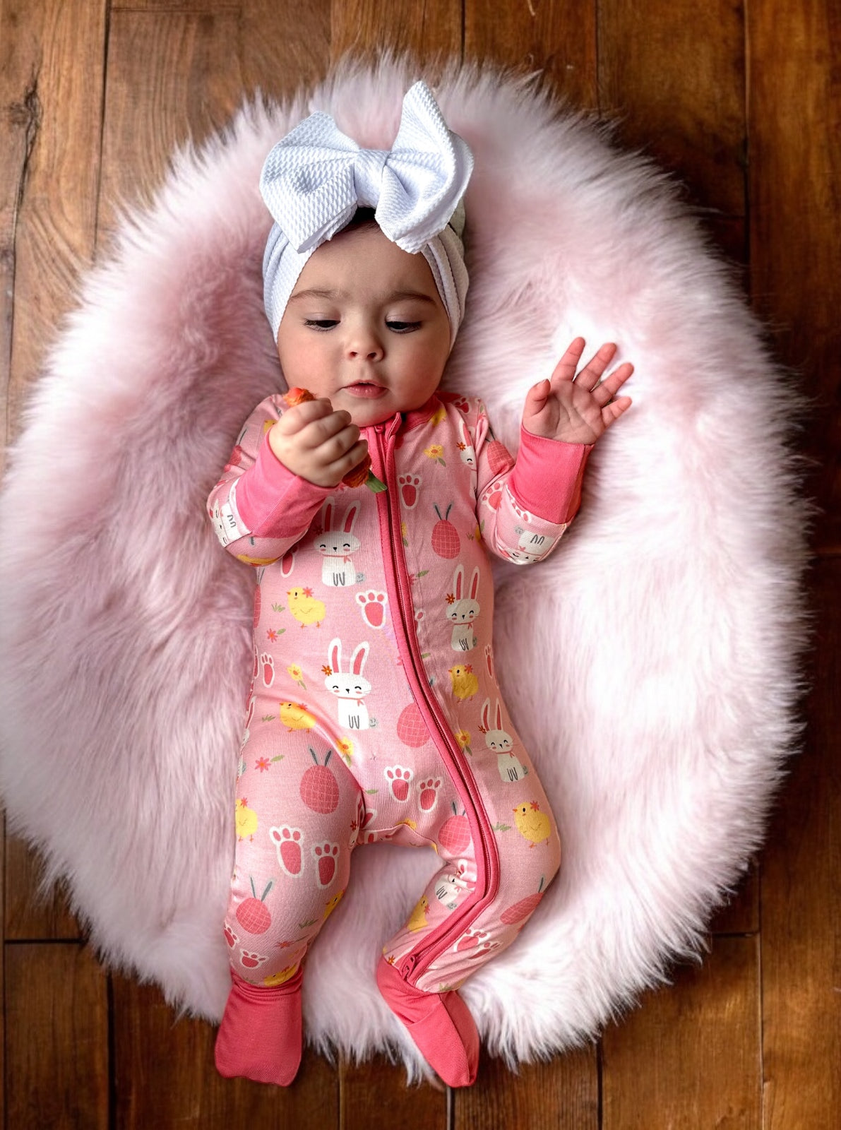 Infant in pink bunny-patterned onesie rests on a fluffy rug, holding a snack and wearing a large white bow.