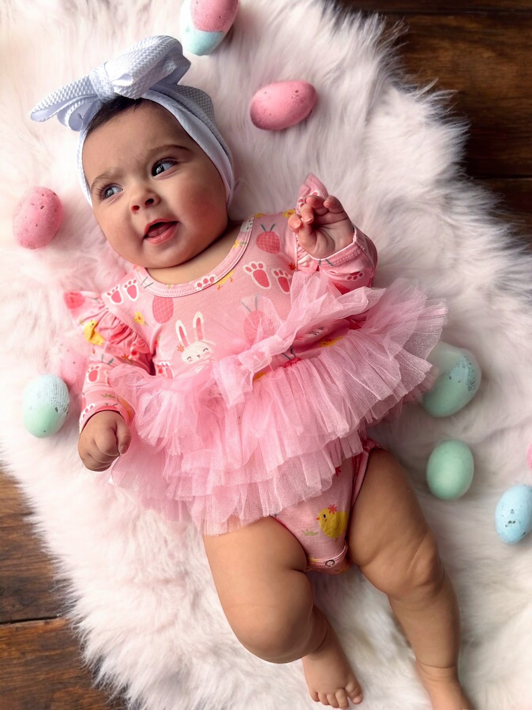 Baby in a pink bunny outfit with pink tulle, surrounded by colorful Easter eggs on a soft, white blanket.