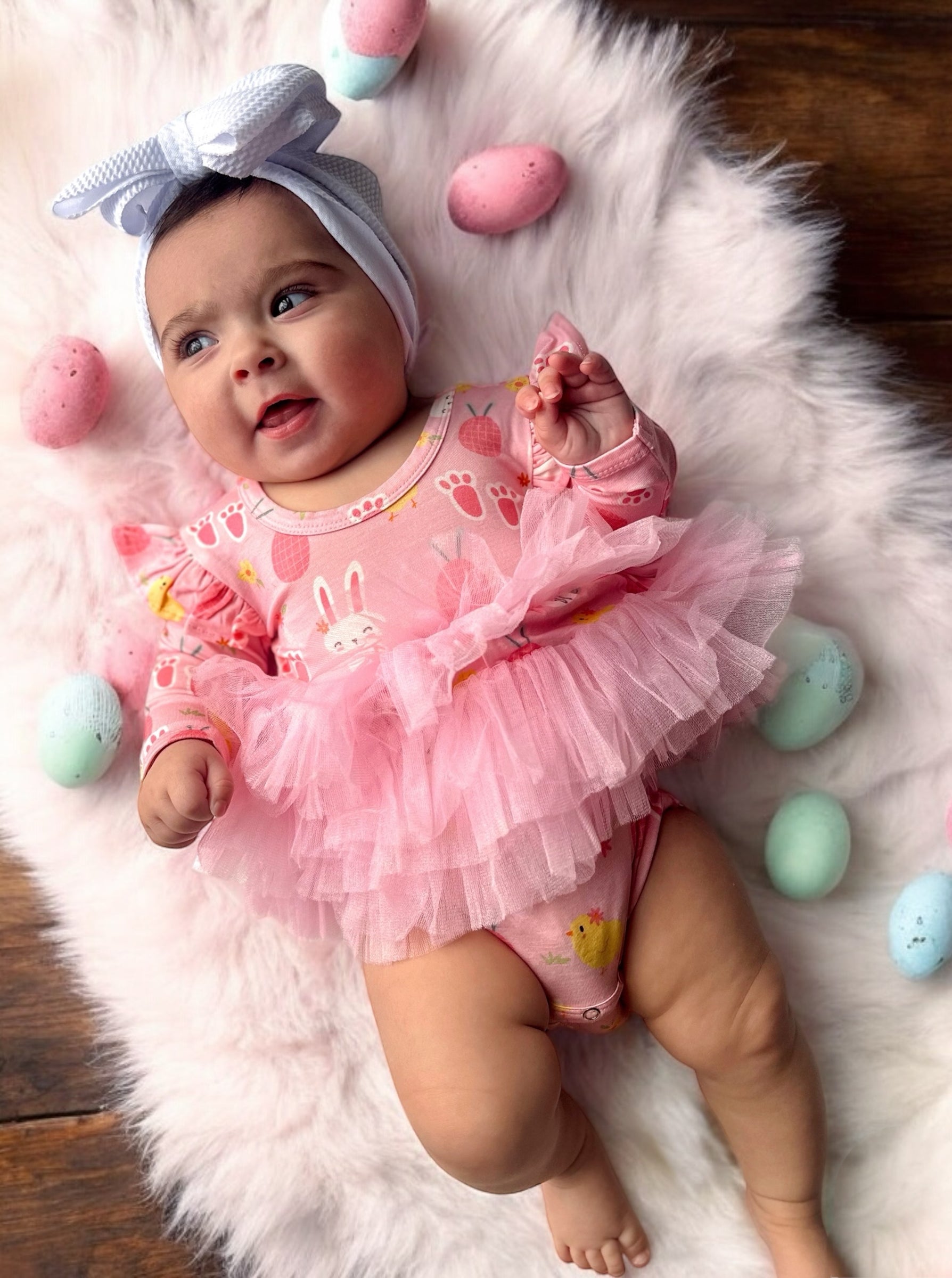 Baby in a pink bunny outfit with pink tulle, surrounded by colorful Easter eggs on a soft, white blanket.