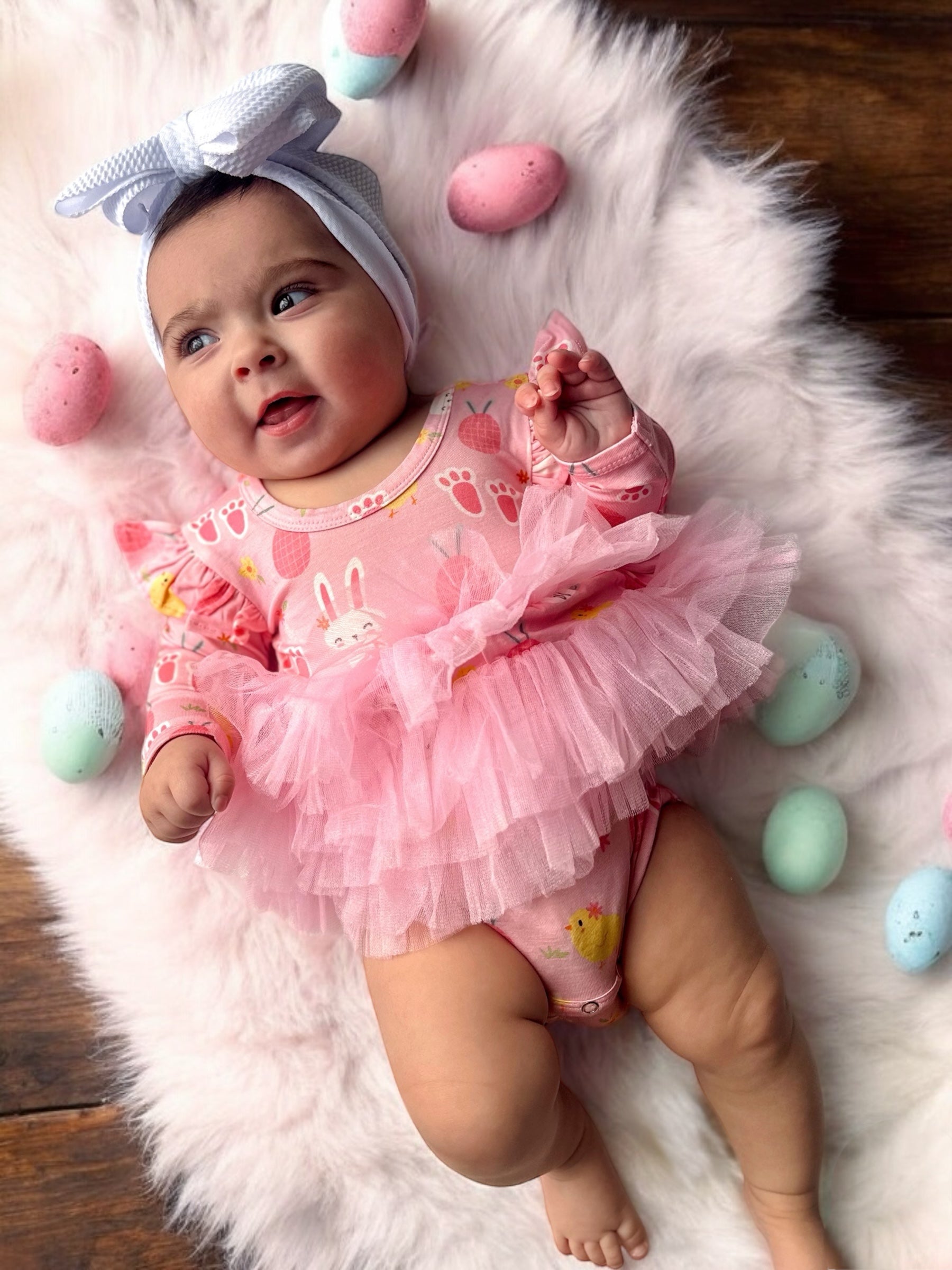 Baby in a pink bunny outfit with pink tulle, surrounded by colorful Easter eggs on a soft, white blanket.