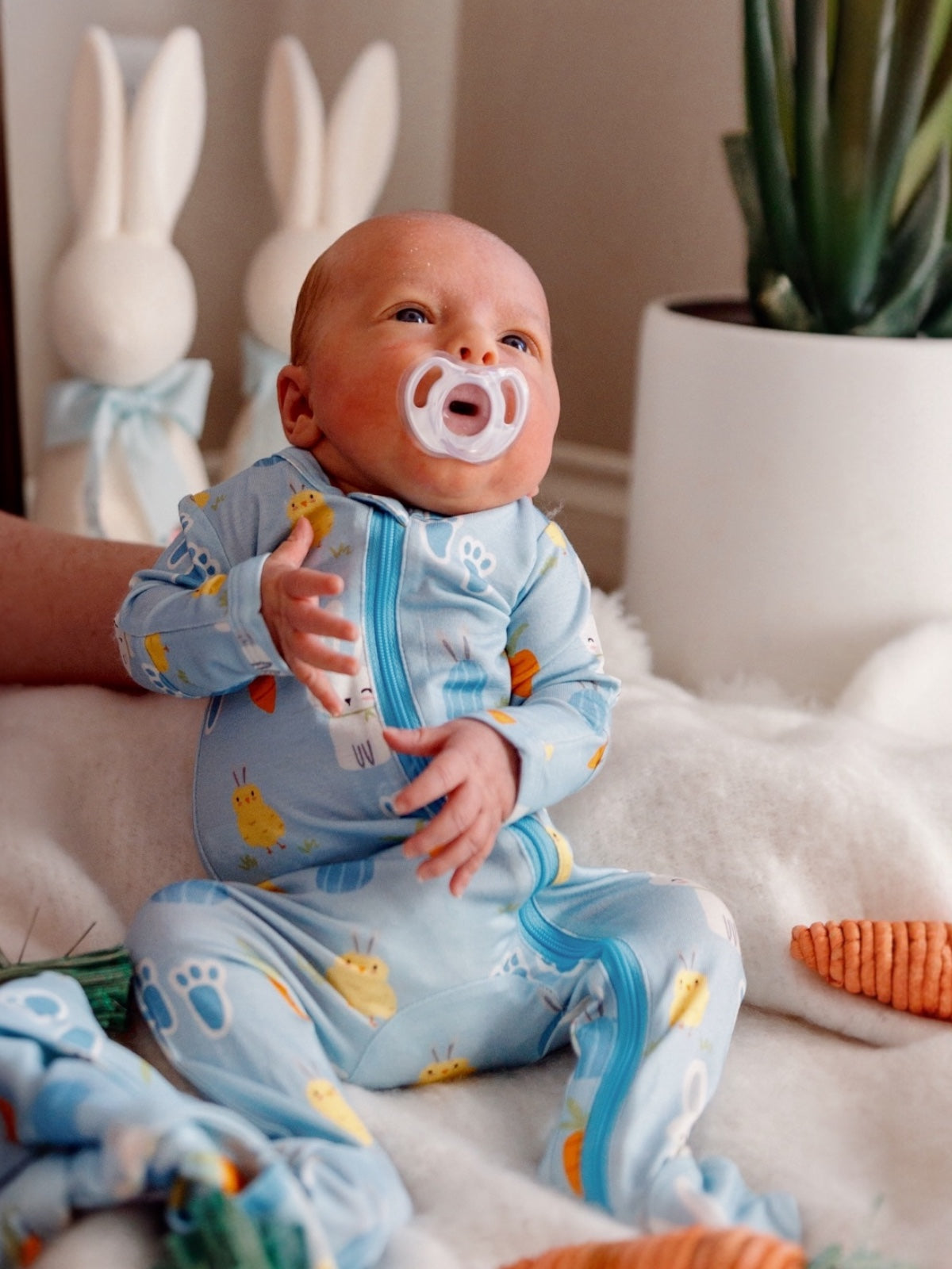 Baby in blue pajamas with animal patterns sits on a blanket, holding a pacifier, surrounded by decorative bunnies.