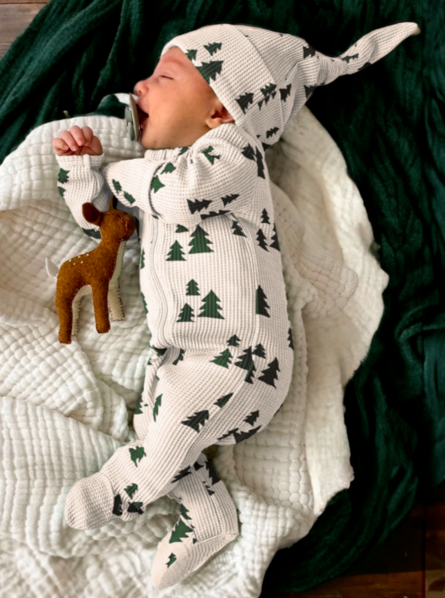 Baby in a patterned outfit with trees, holding a toy deer, lying on a cozy blanket.