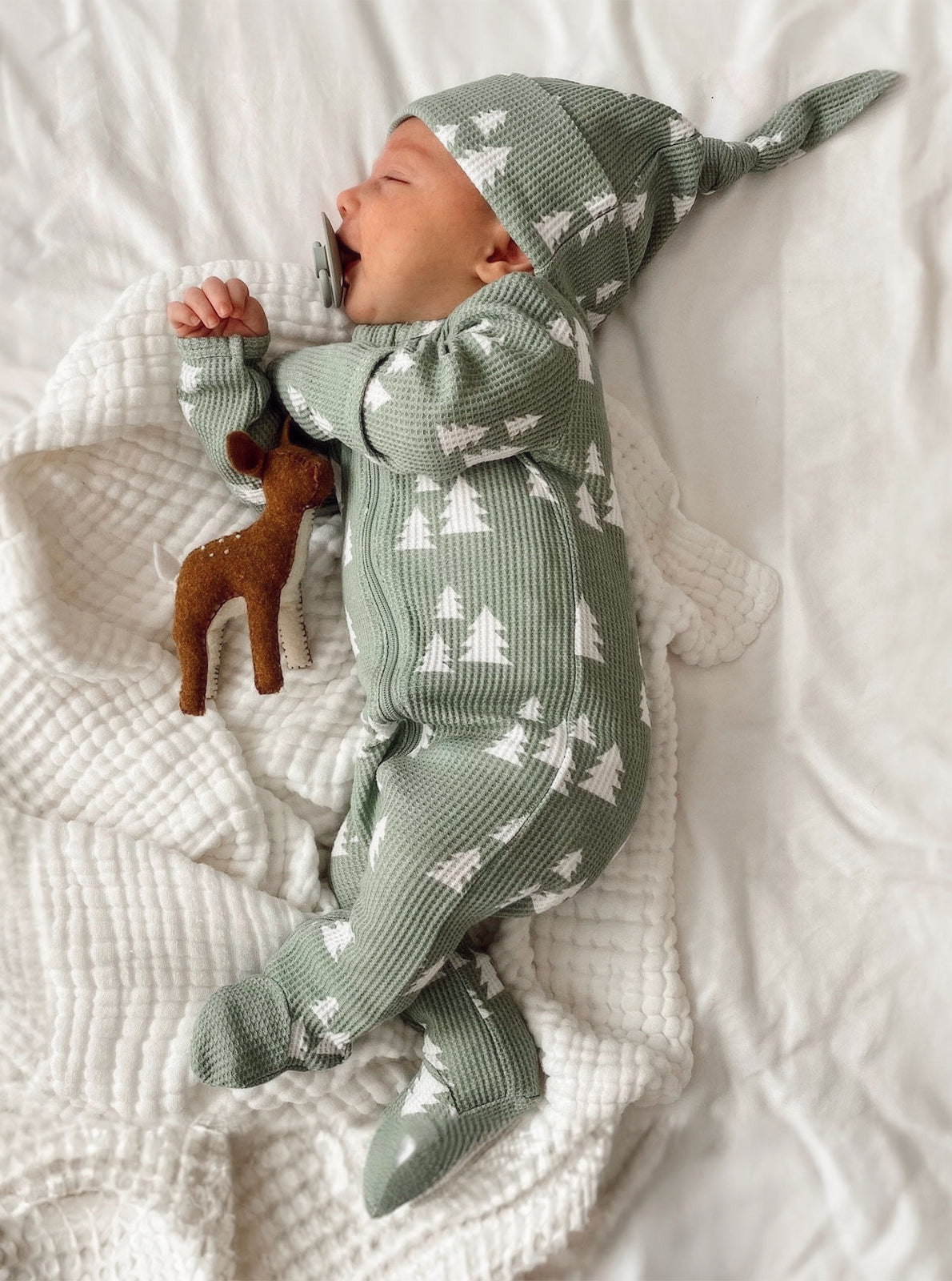 Baby sleeping on a soft blanket, wearing a green patterned outfit and hat, holding a toy deer.