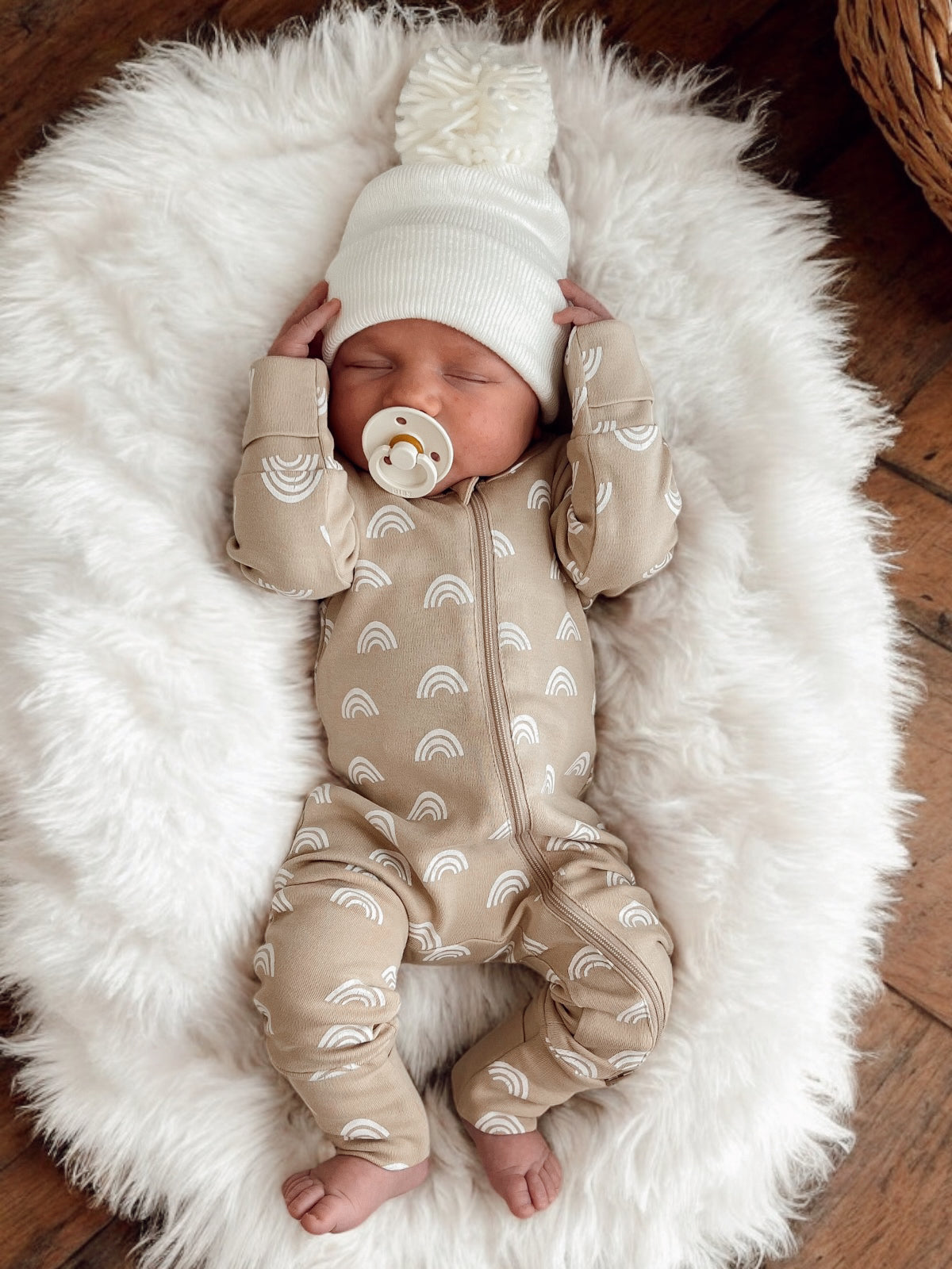 Baby sleeping on a fluffy white blanket, wearing a beige onesie with rainbow patterns and a cozy white hat.