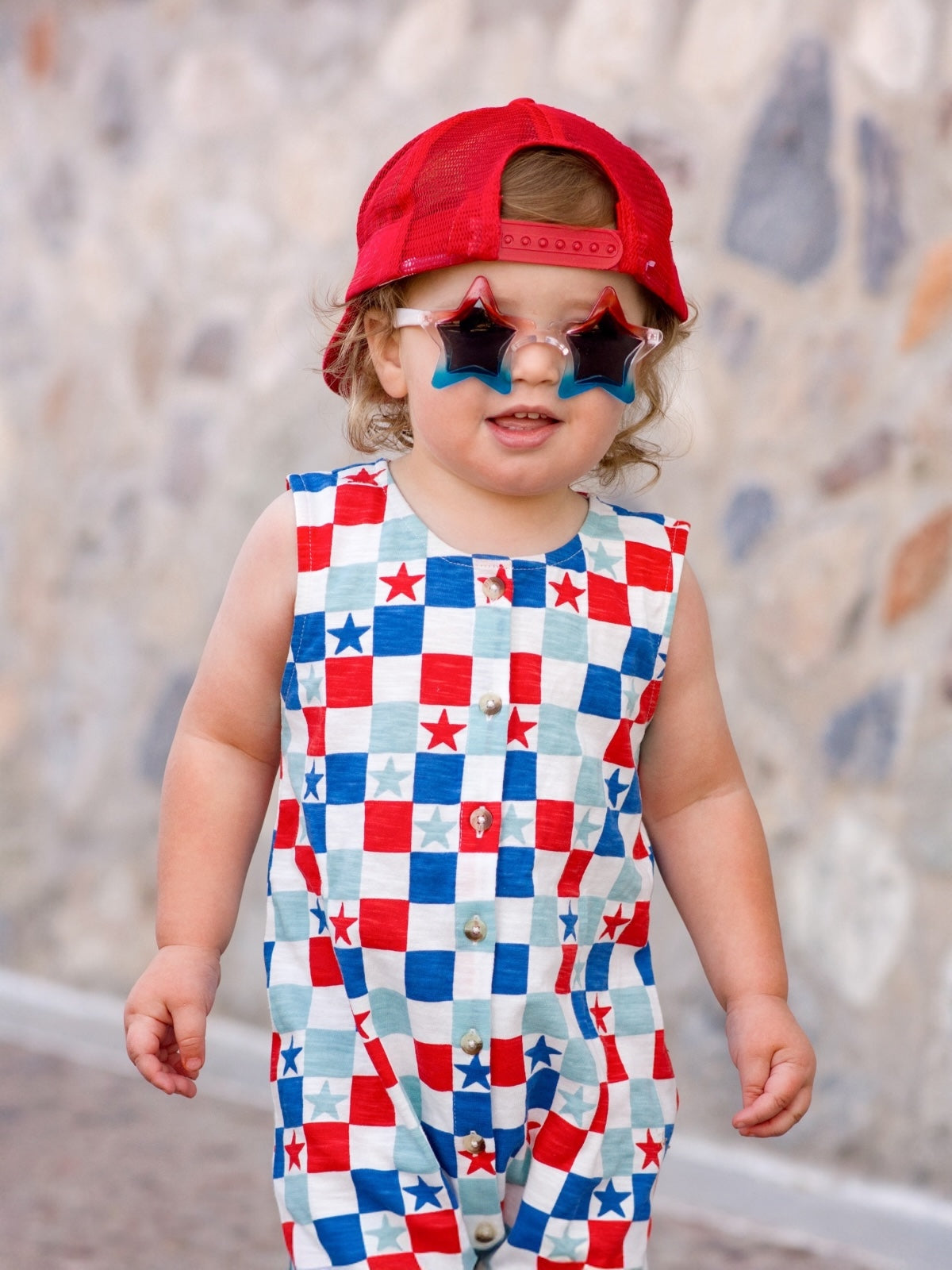 Child in star-shaped sunglasses and a red cap, wearing a colorful plaid jumpsuit, smiles against a stone wall.