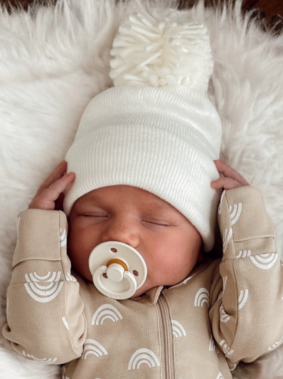 Adorable sleeping baby in a white pom-pom beanie and beige onesie with rainbow patterns on a soft blanket.