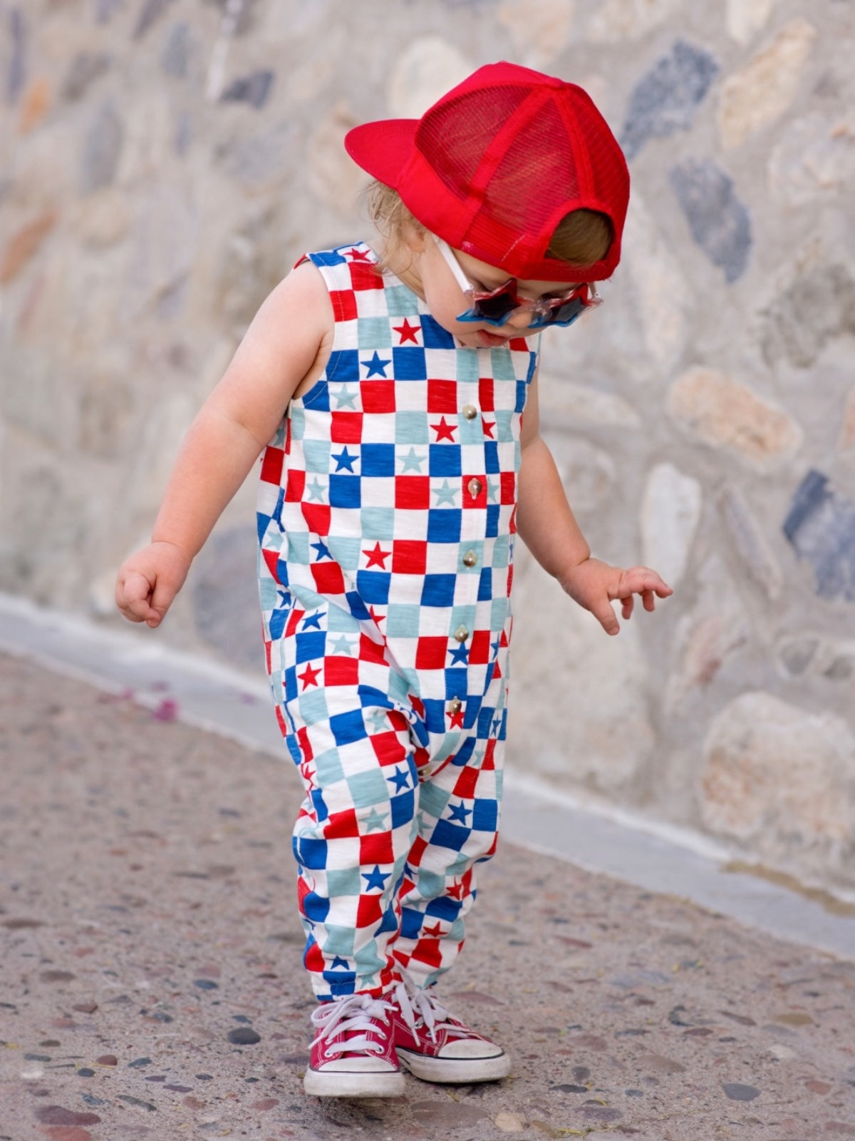 Child in colorful star-patterned outfit and red cap, standing on a stone path, looking down.