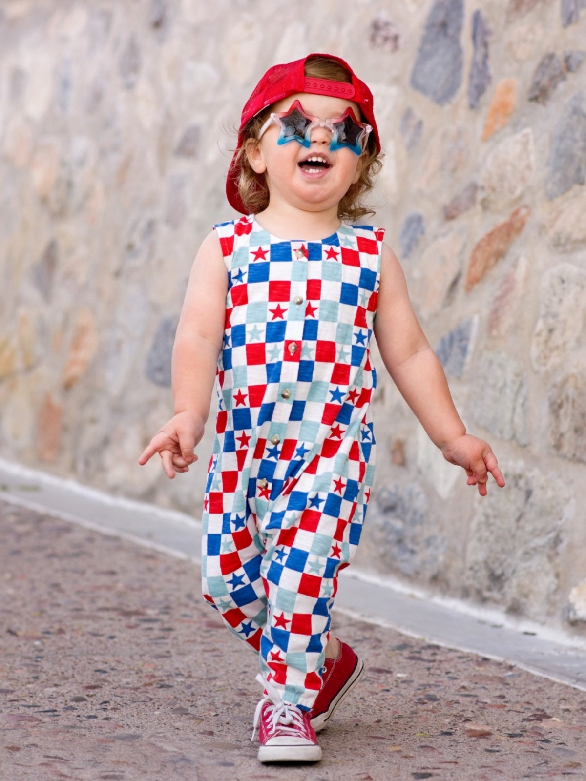 Child in checkerboard outfit with stars, red cap, and star-shaped glasses, walking on a stone path.