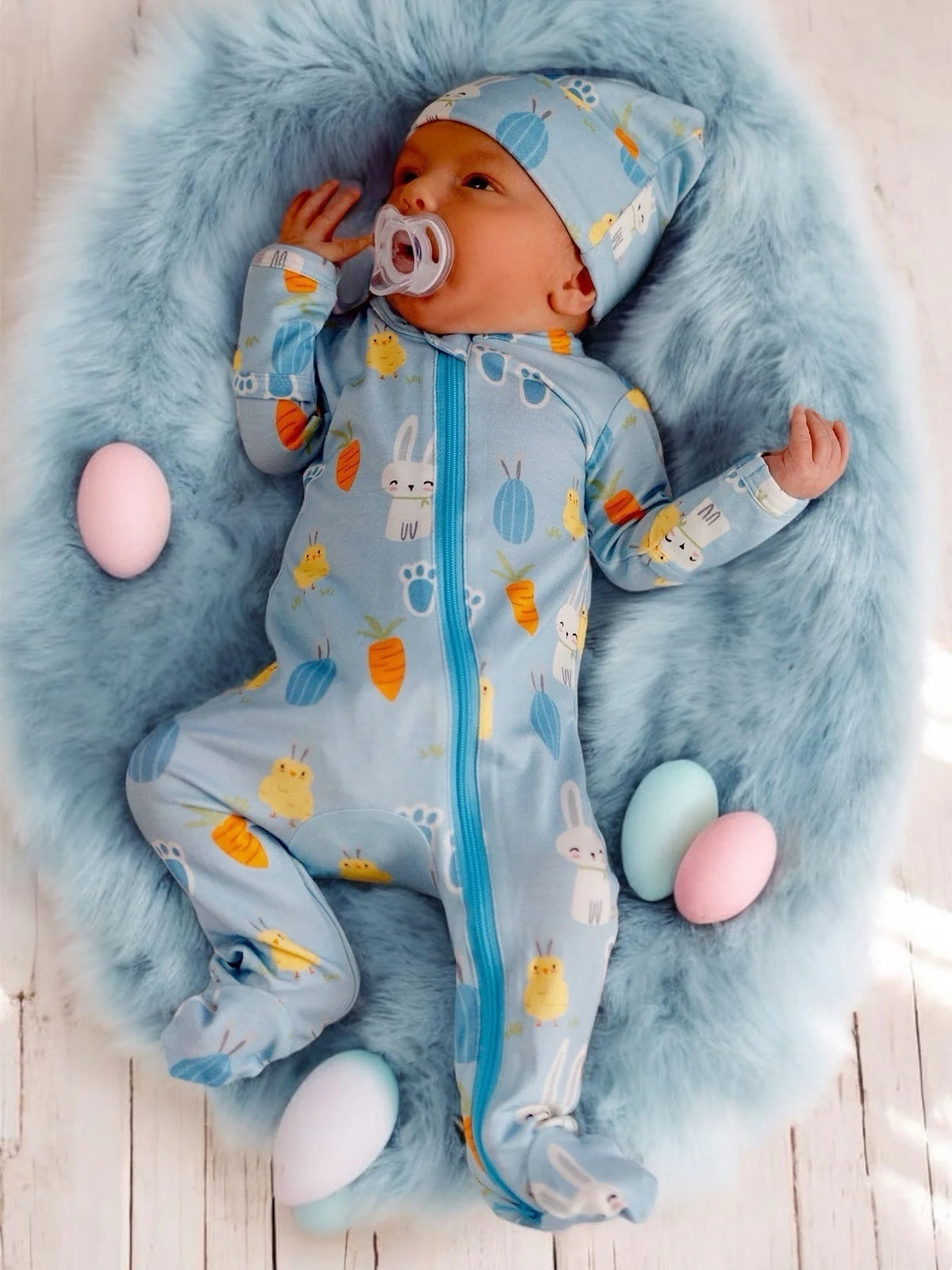 Infant in blue printed pajamas with a pacifier, surrounded by pastel-colored eggs on soft, fluffy bedding.